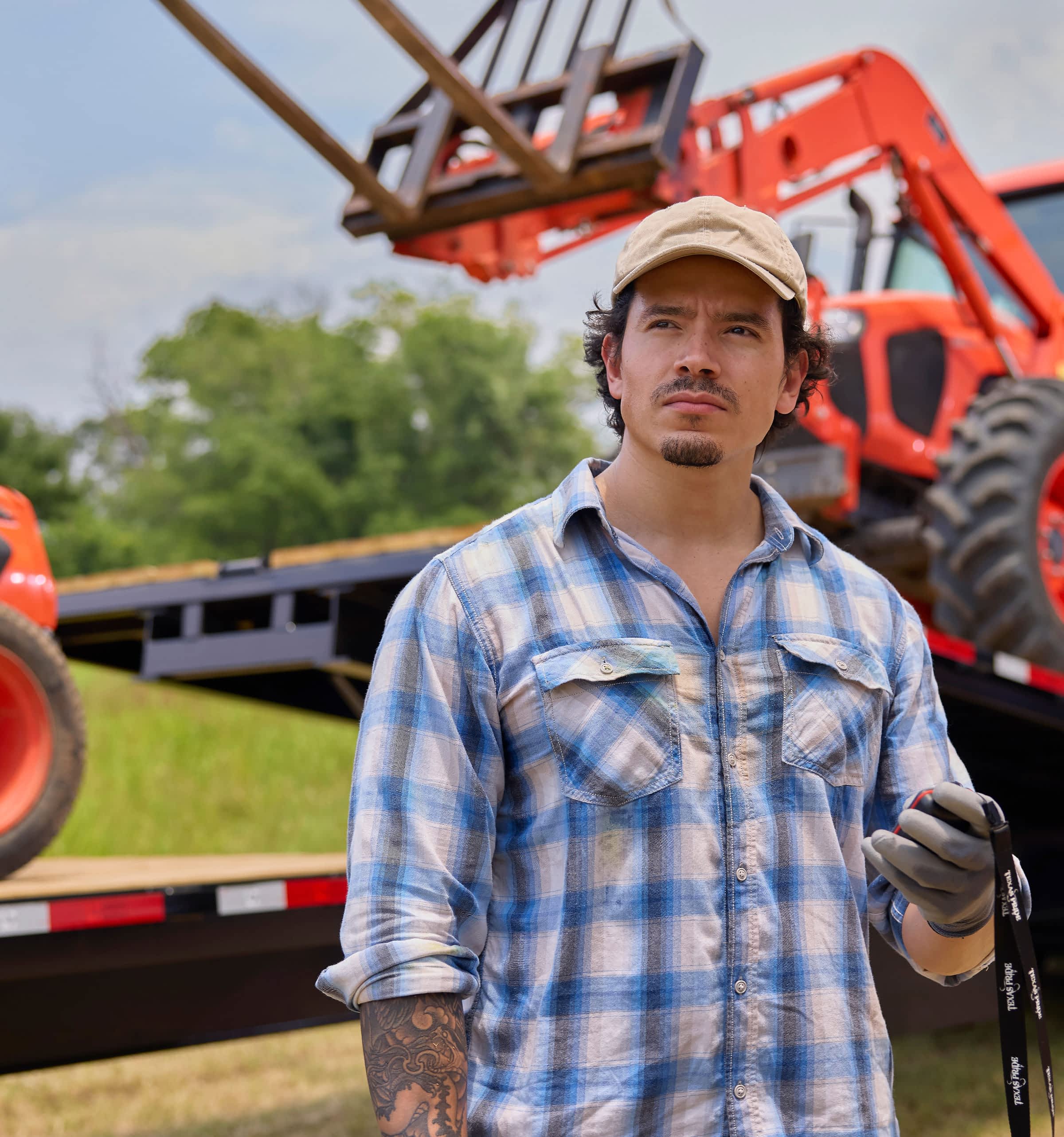 Man in a flannel shirt and baseball cap working outdoors in front of agricultural trailer.