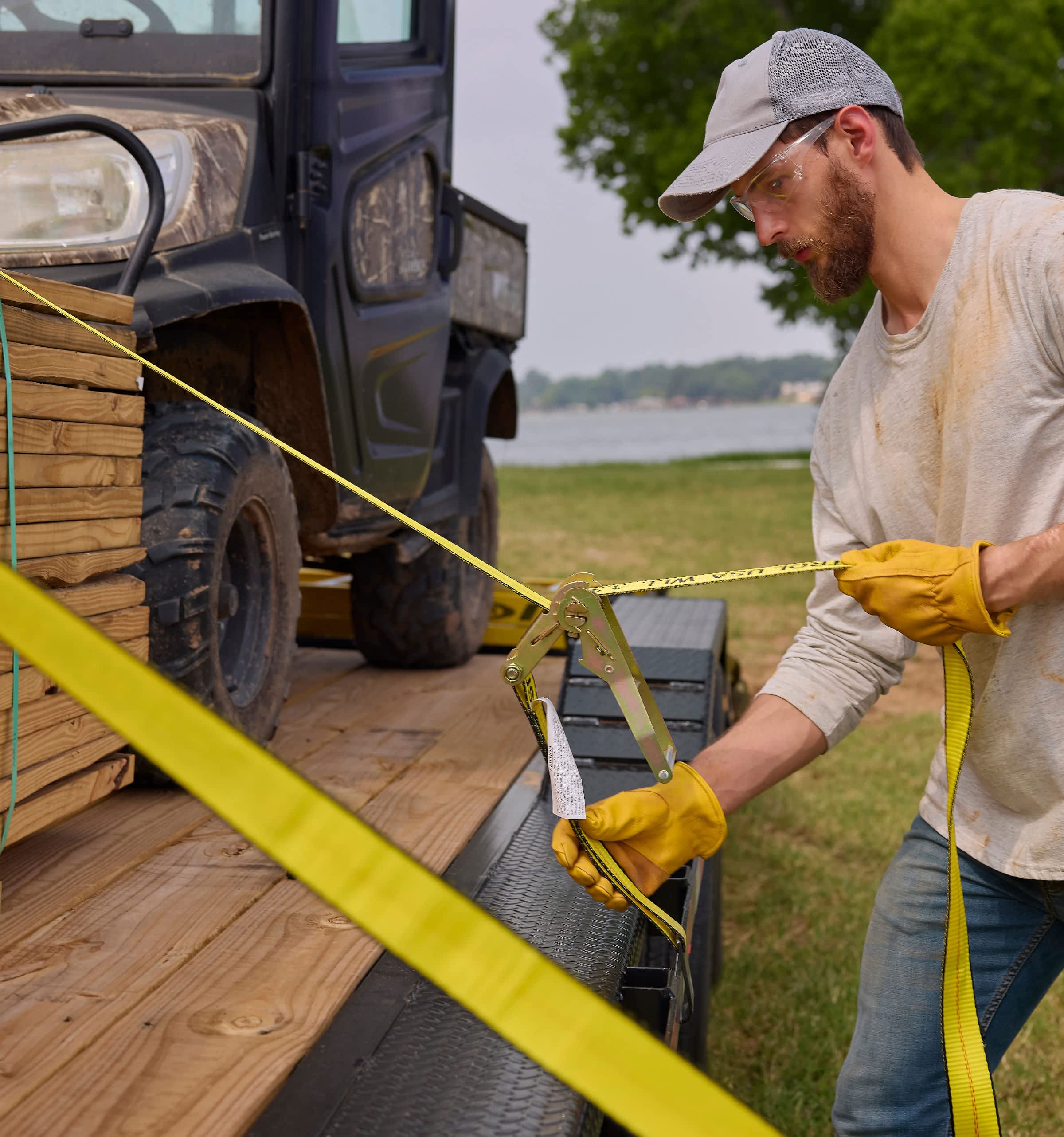 A man in work gloves securing a load of lumber and a vehicle on a trailer.