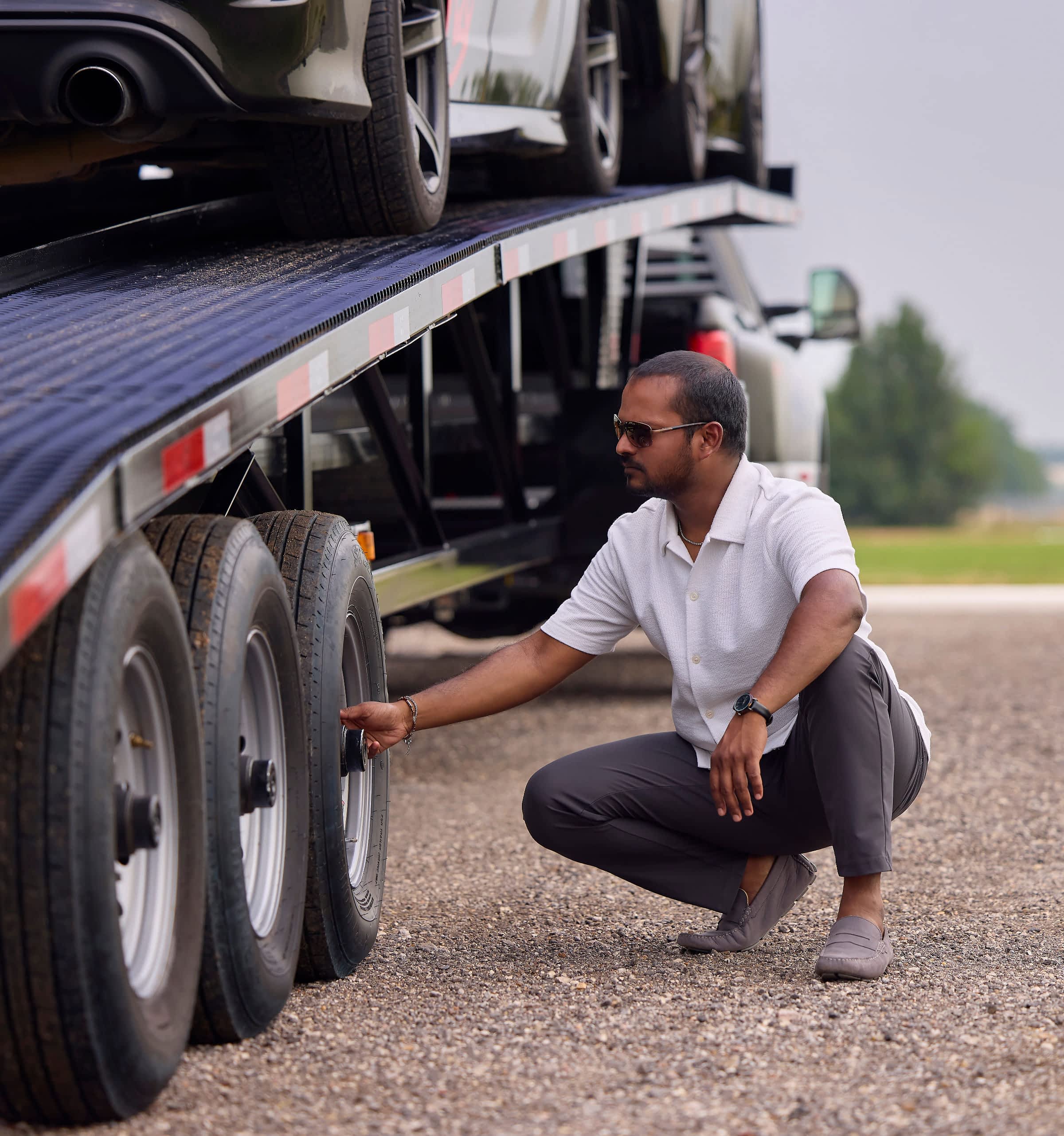A man in sunglasses and sharp outfit inspecting a tire trailer loaded up with vehicles.