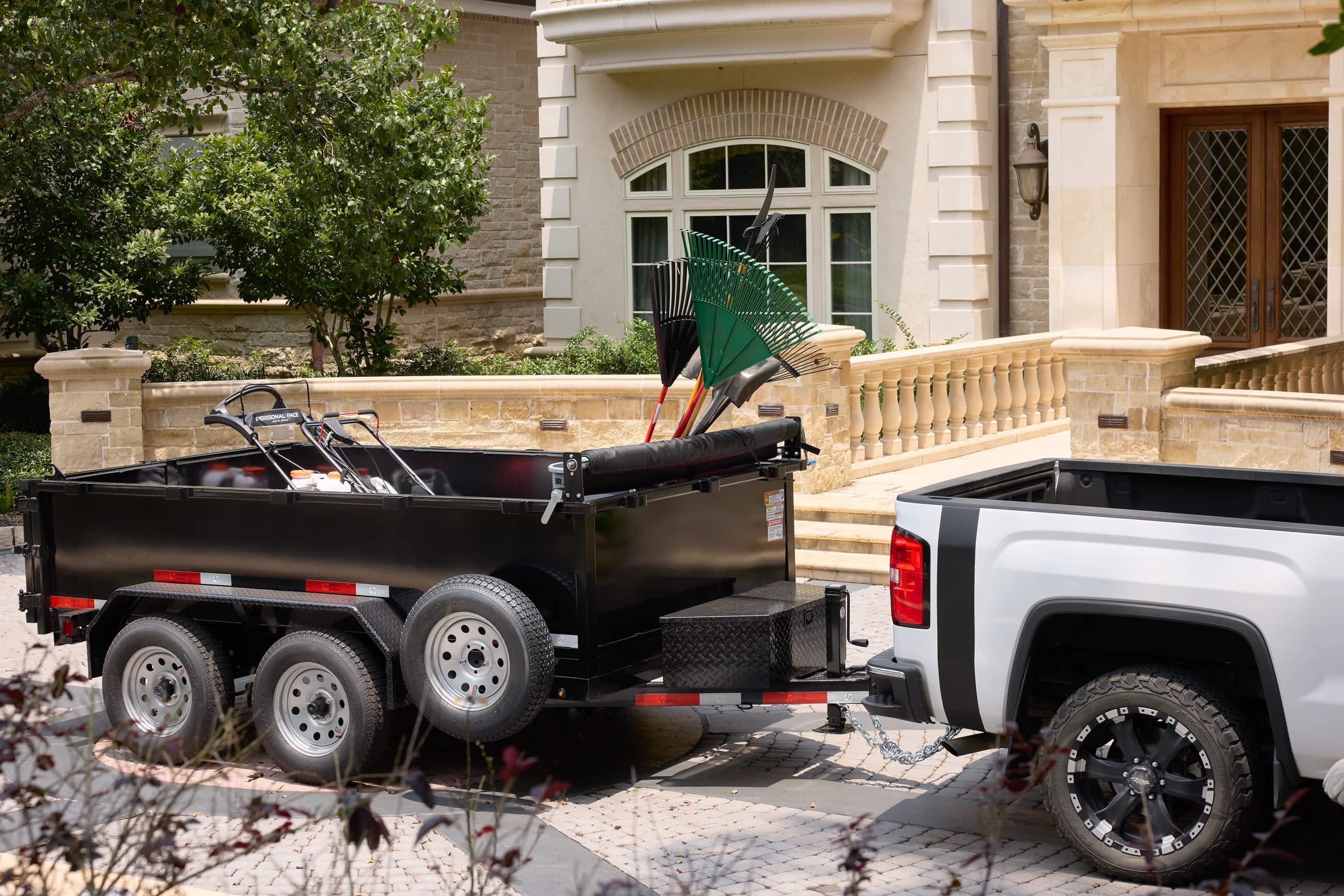 Texas Pride bumper pull dump trailer with tandem axles and mounted toolbox, loaded with landscaping equipment including rakes and lawn tools, hitched to a white pickup truck in front of a residential jobsite.