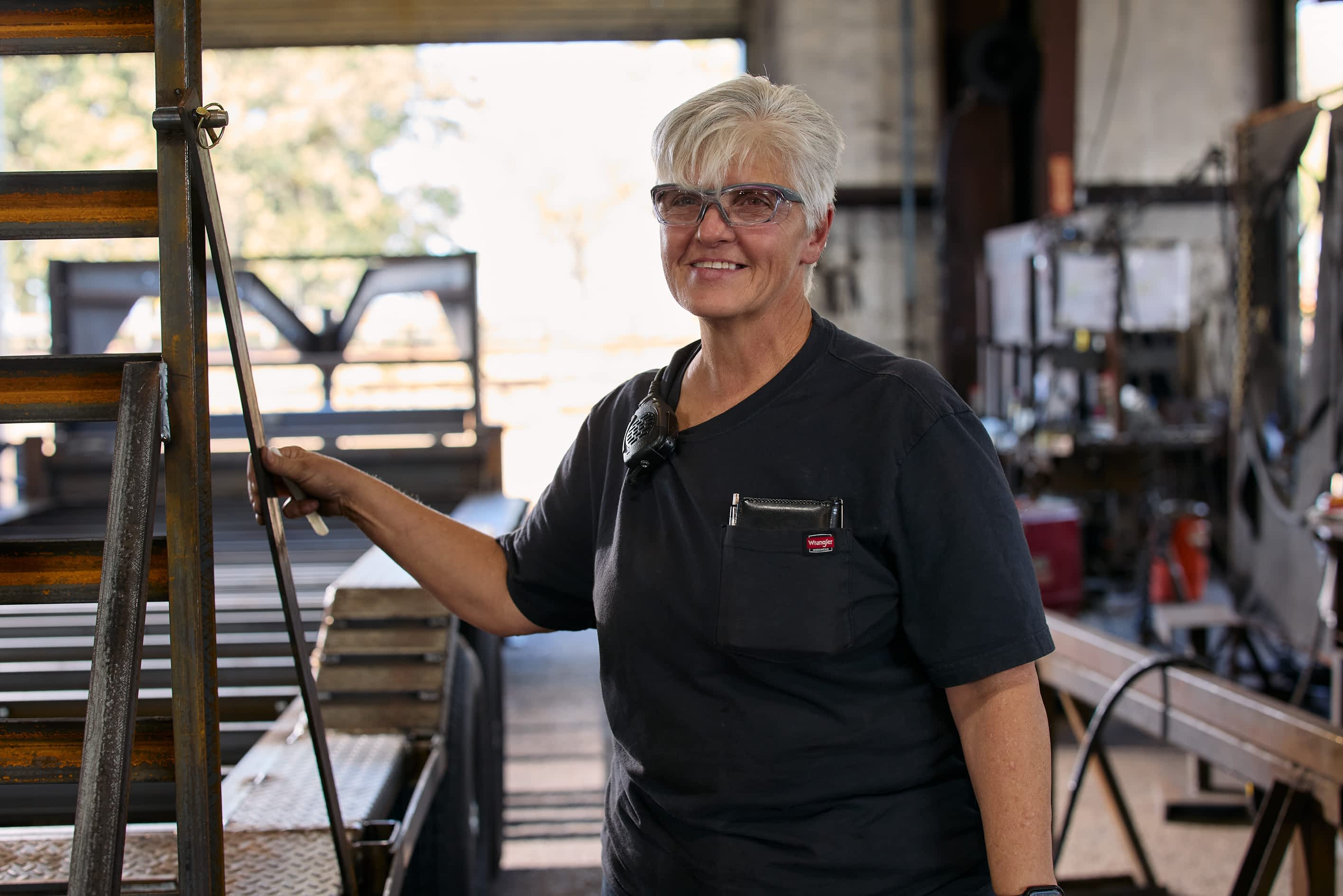 A woman with short grey hair smiles at the camera, standing next to metal structures in a Texas Pride Trailers manufacturing facility.