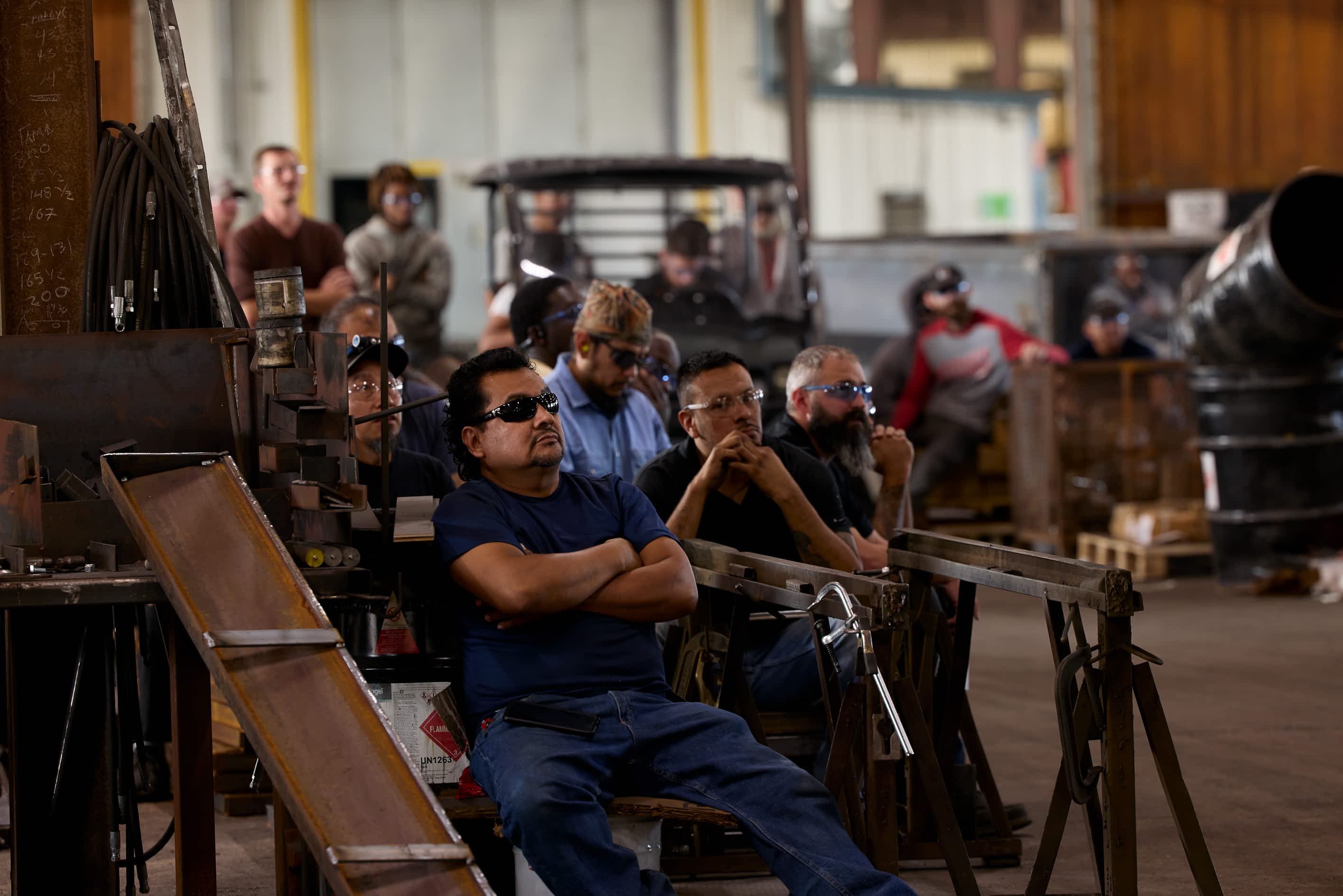 A group of workers sit on trailer components in a Texas Pride Trailers manufacturing facility, listening to a speaker.