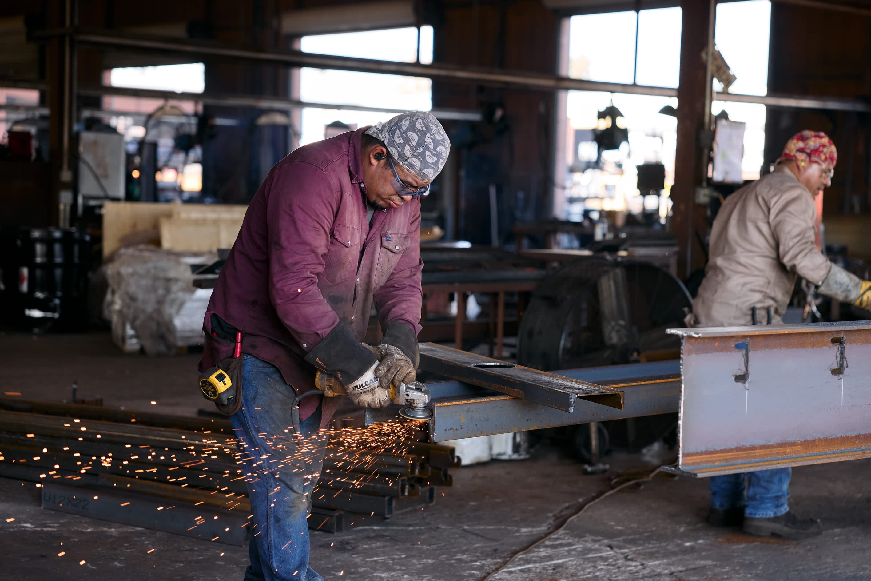 A worker uses an angle grinder on a metal beam, creating sparks, as part of Texas Pride Trailers manufacturing in a factory. Another worker is in the background.