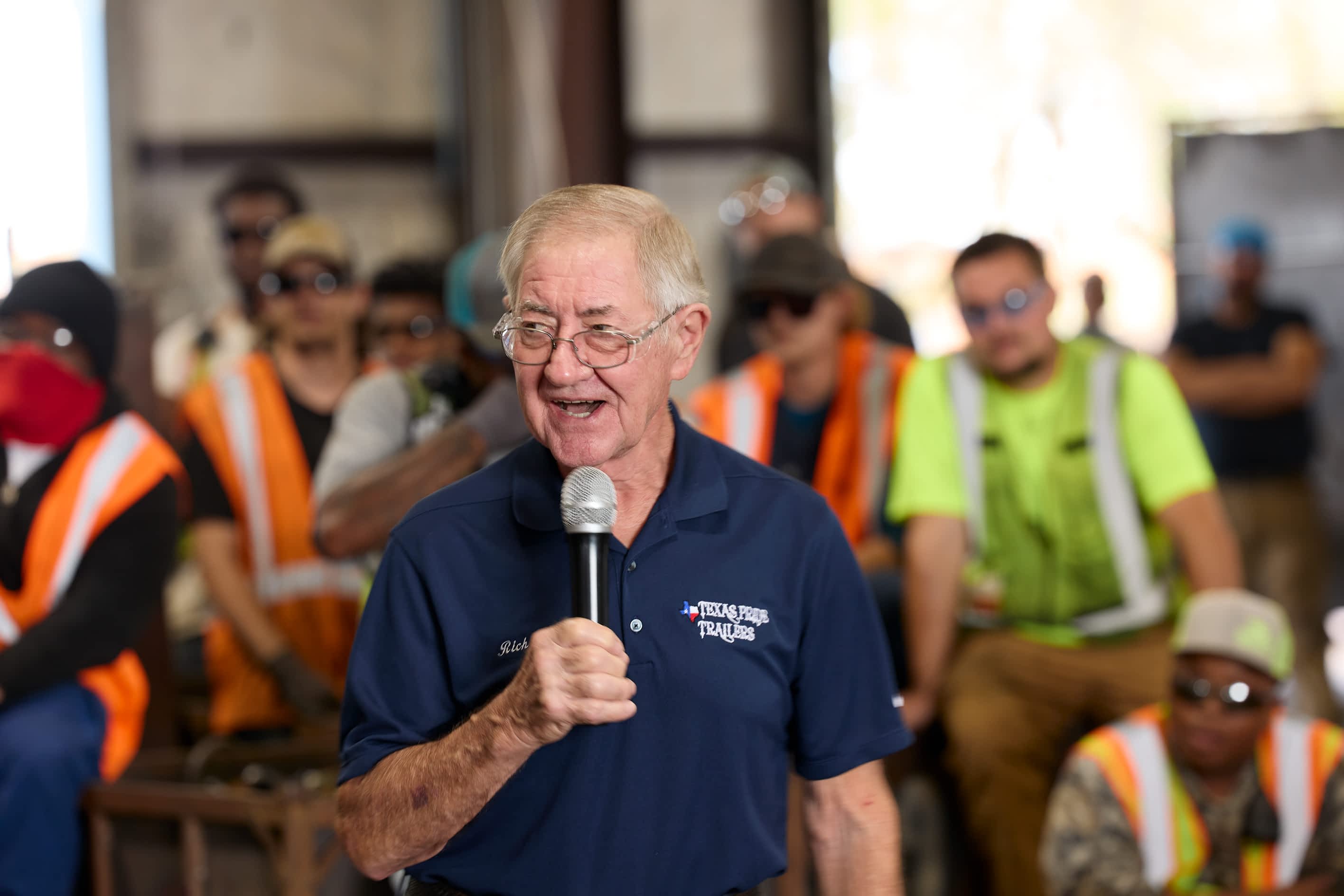 A man in a Texas Pride Trailers logo shirt speaks into a microphone in a factory setting with a crowd of people listening in the background.