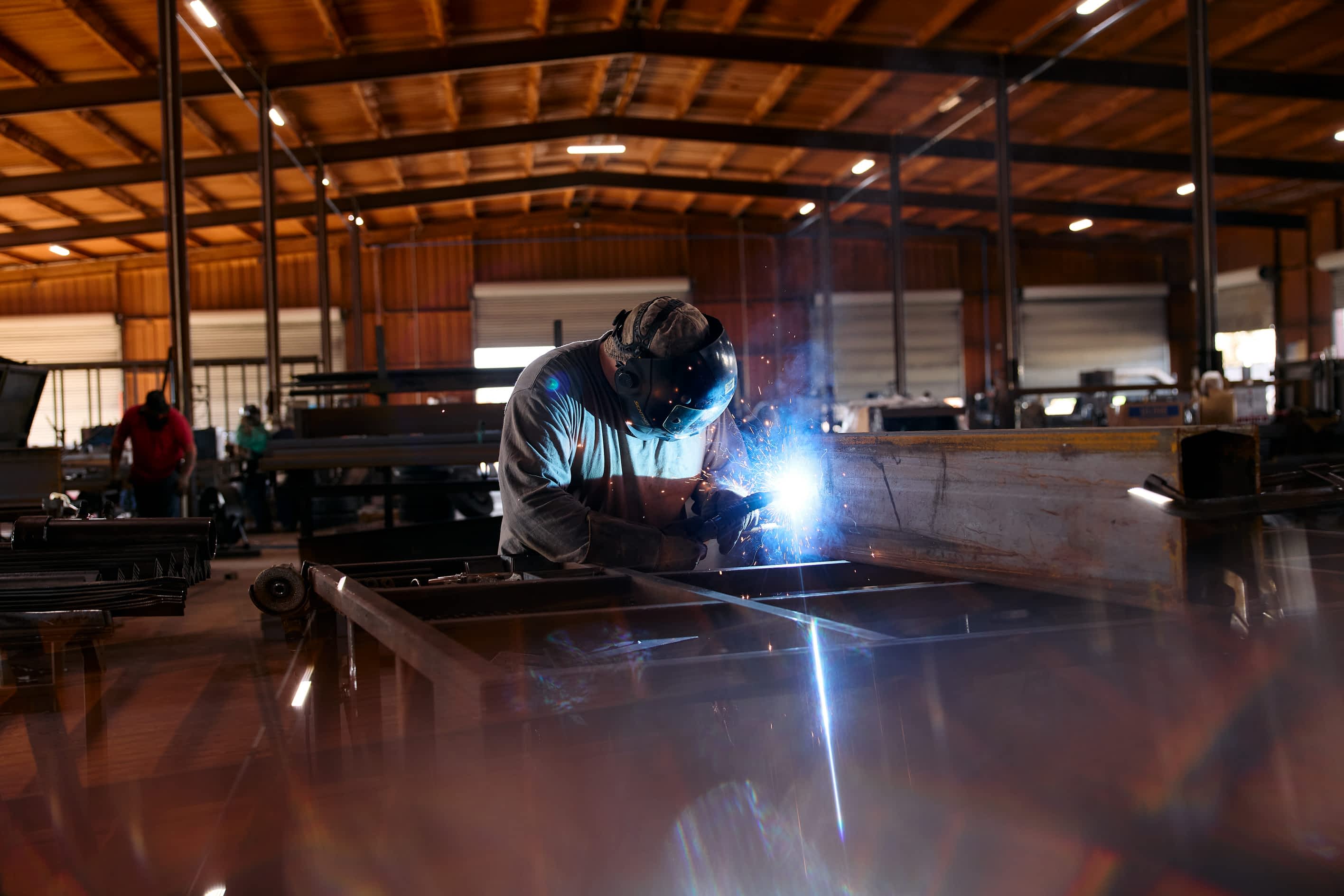 A worker wearing a welding helmet welds a Texas Pride Trailers frame in a factory. Welding light and sparks are visible.