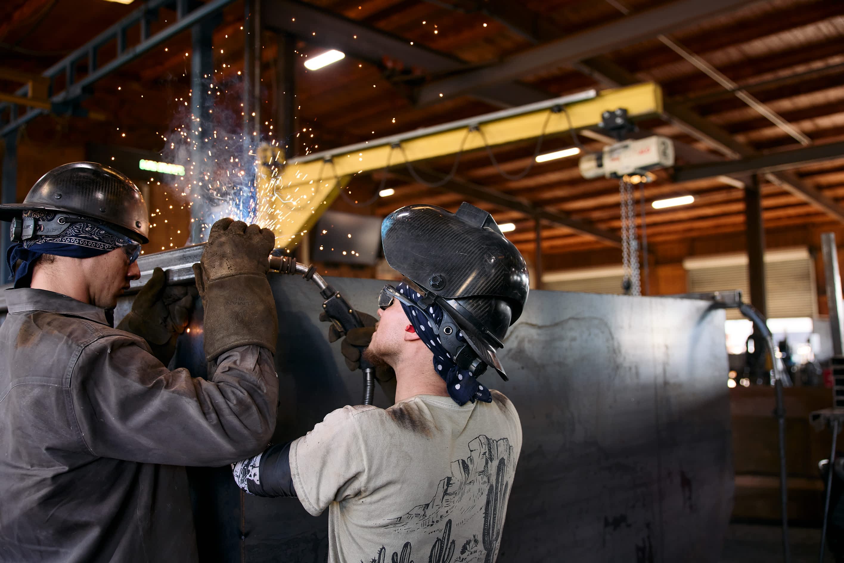 Two workers in welding helmets are welding a Texas Pride Trailers component in a factory, creating bright sparks and light.