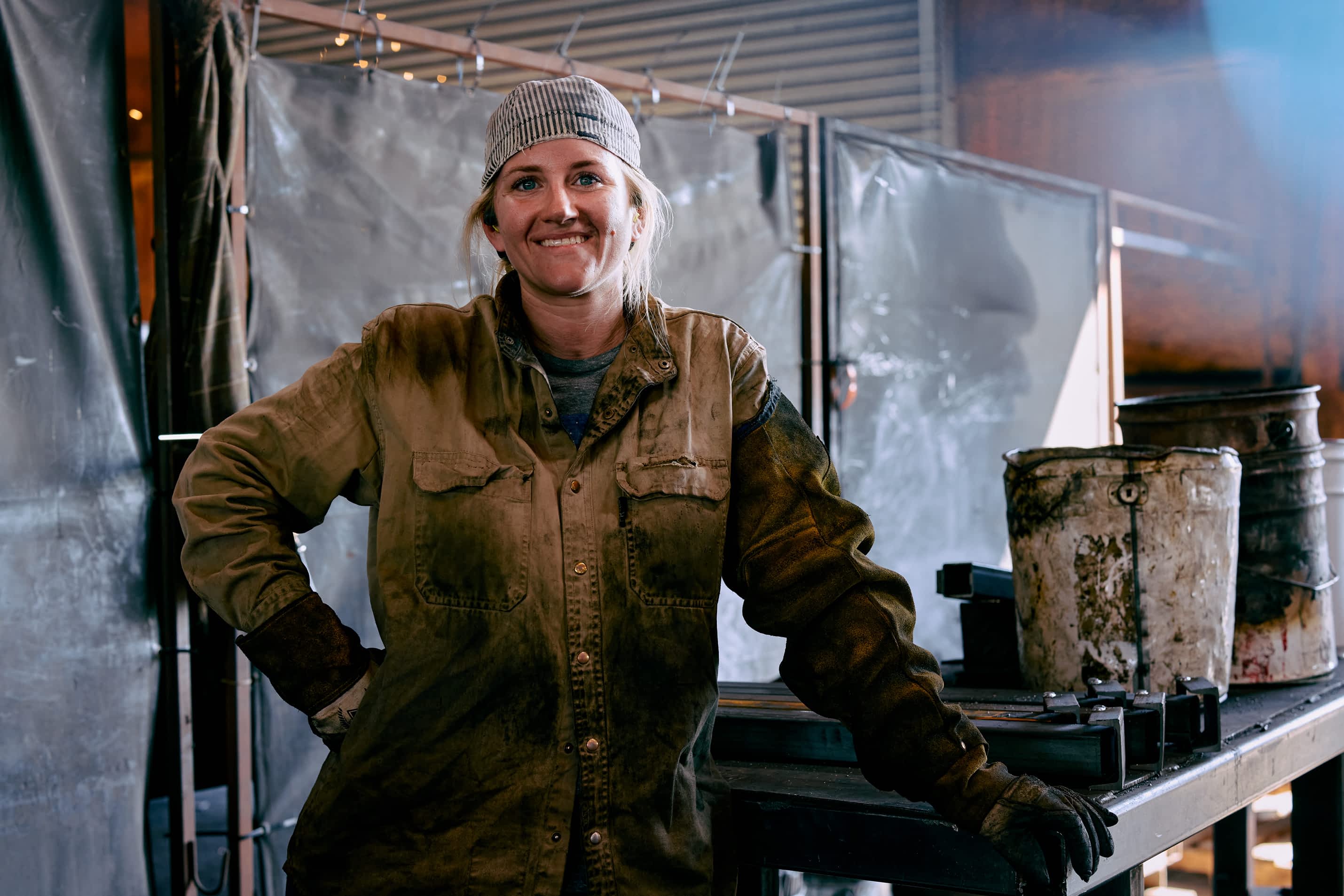 A worker in a striped cap, worn work shirt, and gloves leans against a metal structure and smiles in a Texas Pride Trailers manufacturing facility.