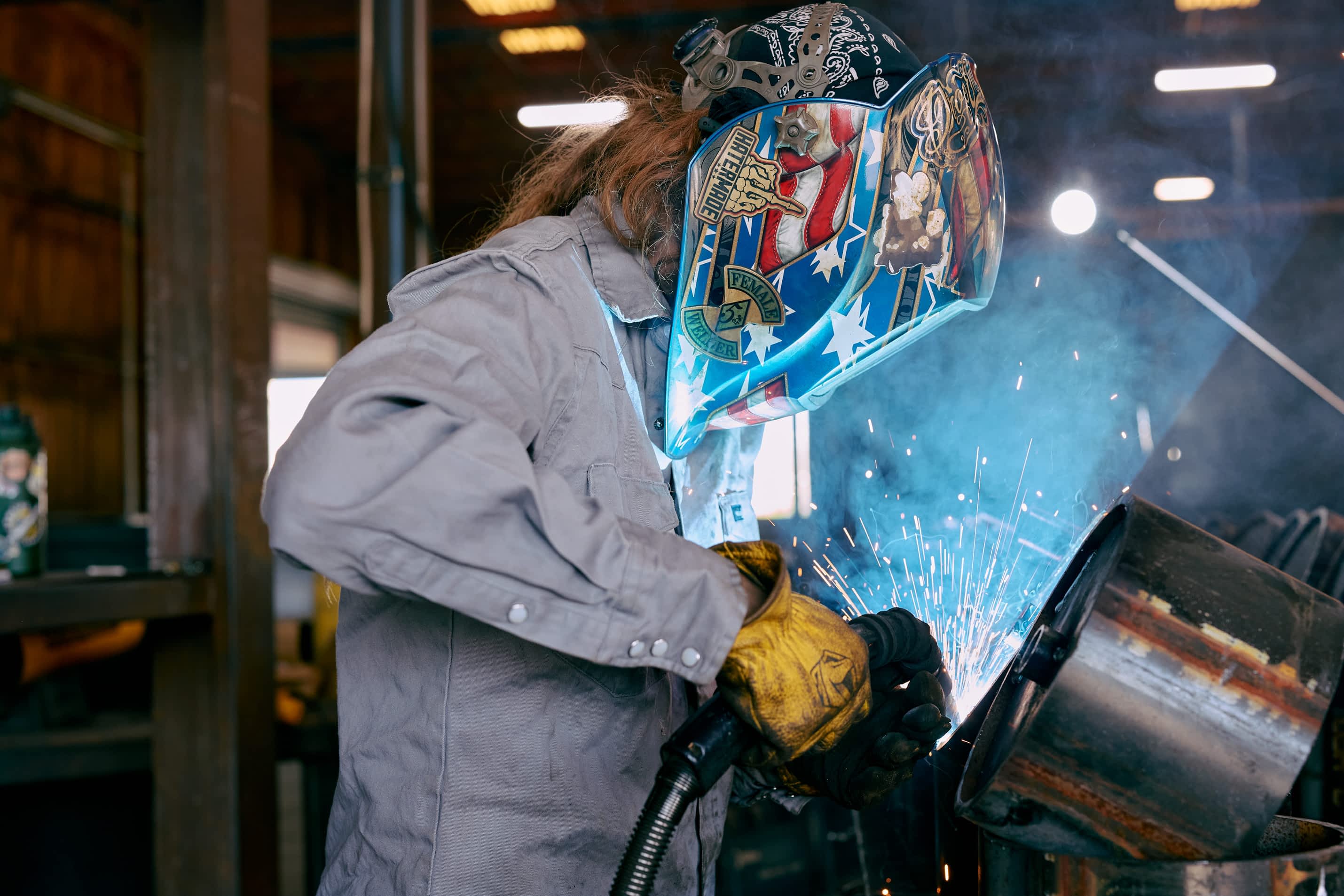 A worker in a decorative welding helmet and gloves is welding a Texas Pride Trailers component in a factory. Sparks and smoke are visible.