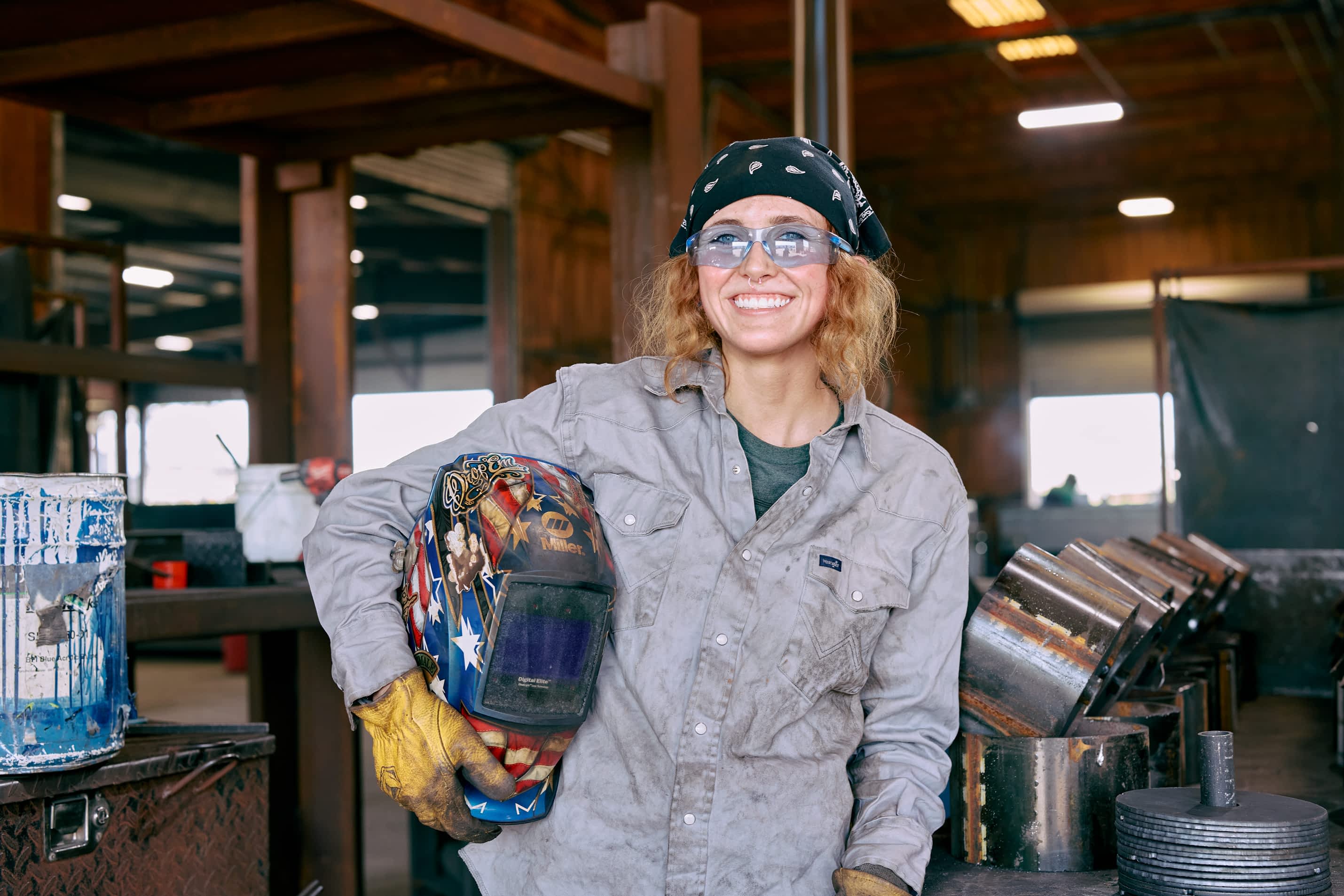A female worker wearing a bandana and safety glasses smiles at the camera, holding a welding helmet in a Texas Pride Trailers manufacturing facility.