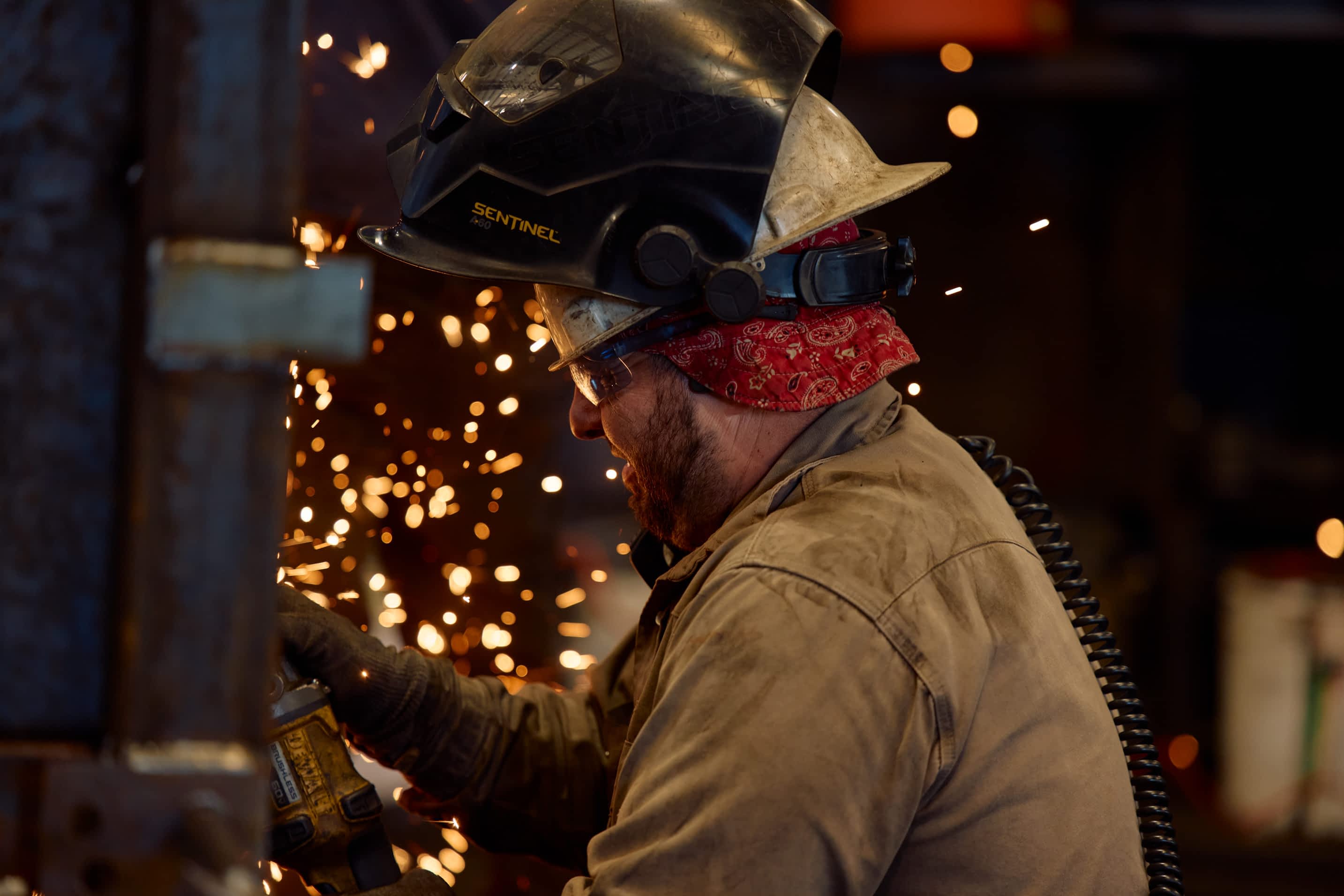 A worker wearing a welding helmet and safety glasses uses an angle grinder, creating many sparks, on a Texas Pride Trailers component in a factory.