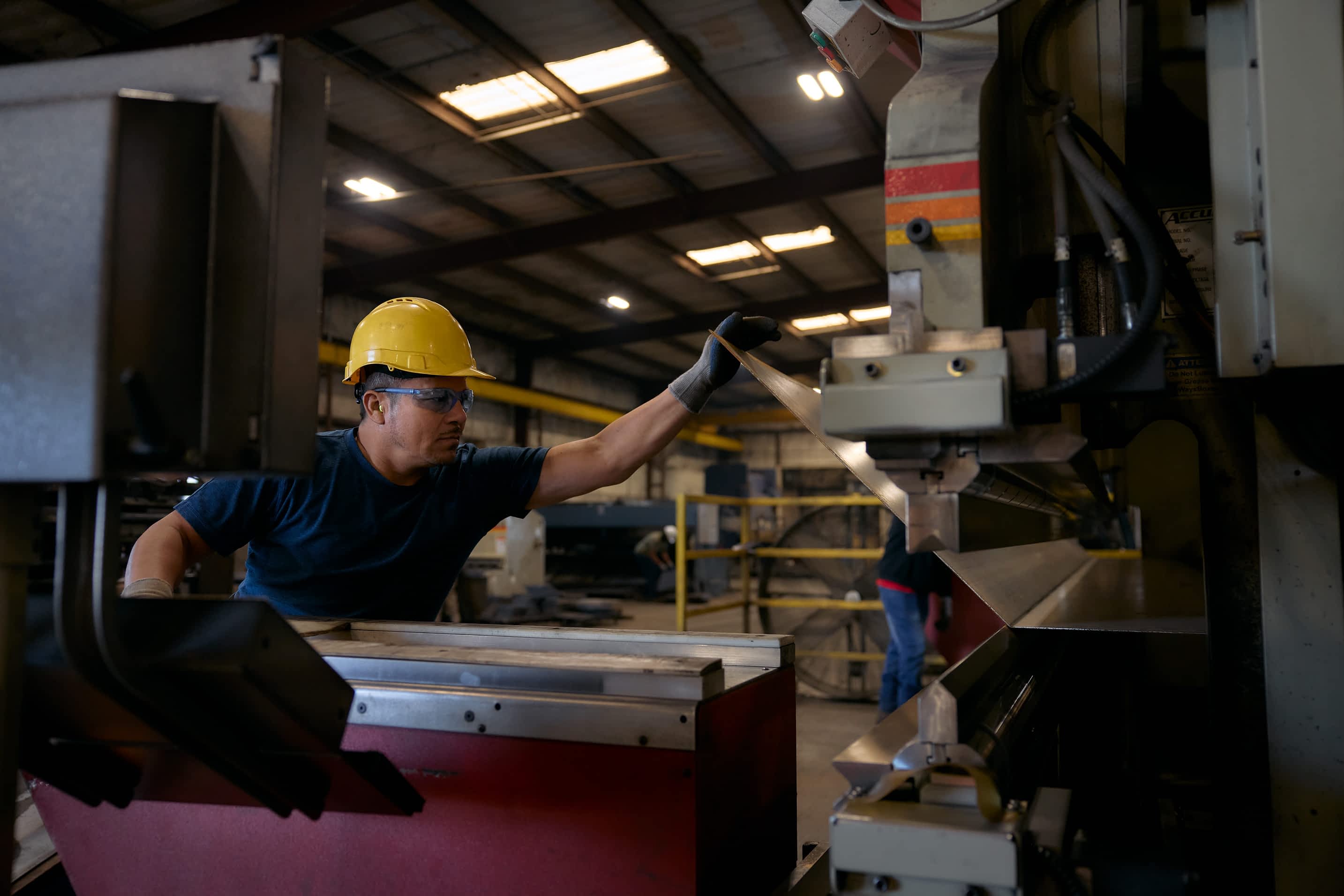 A worker in a hard hat and gloves operates a press machine on a metal piece for a Texas Pride Trailers component in a factory.