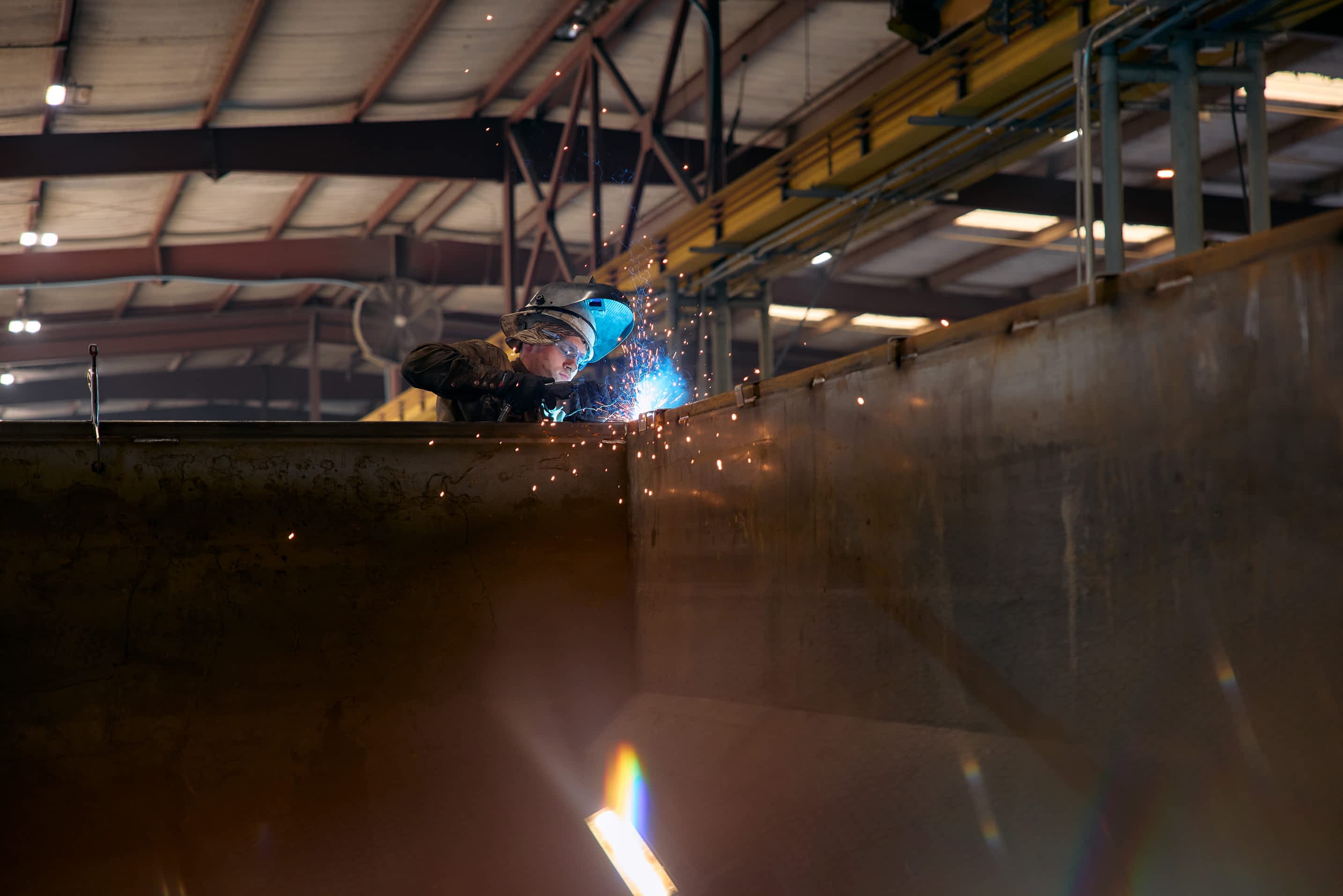 A worker wearing a welding helmet is welding a large metal structure for a Texas Pride Trailers component in a factory, creating bright sparks.