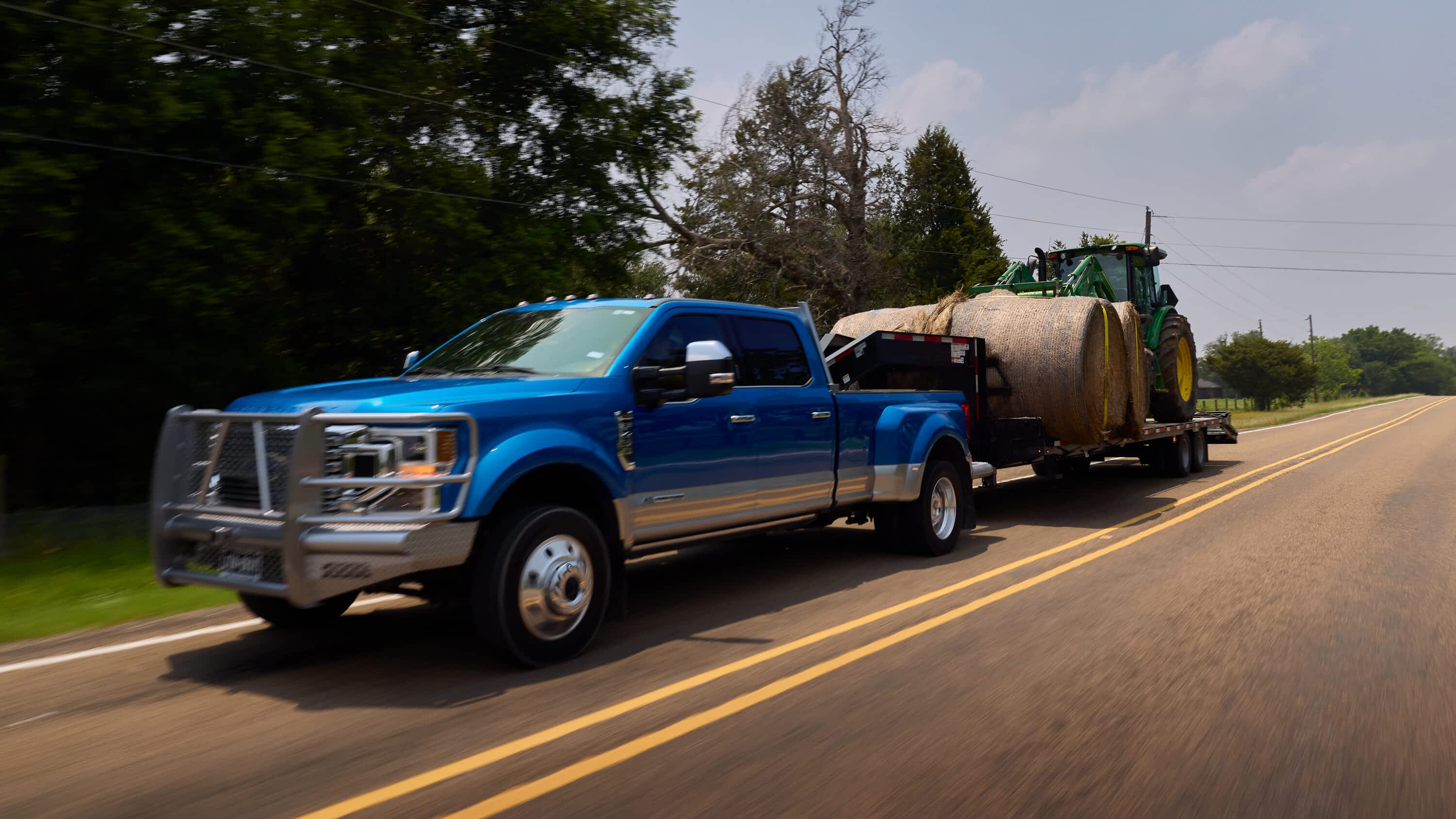 A blue dually pickup truck hauls a Texas Pride power tilt trailer loaded with a green tractor and large hay bales down a rural highway. The trailer's heavy-duty design and low-angle deck demonstrate its hauling capability for farm equipment on the move.