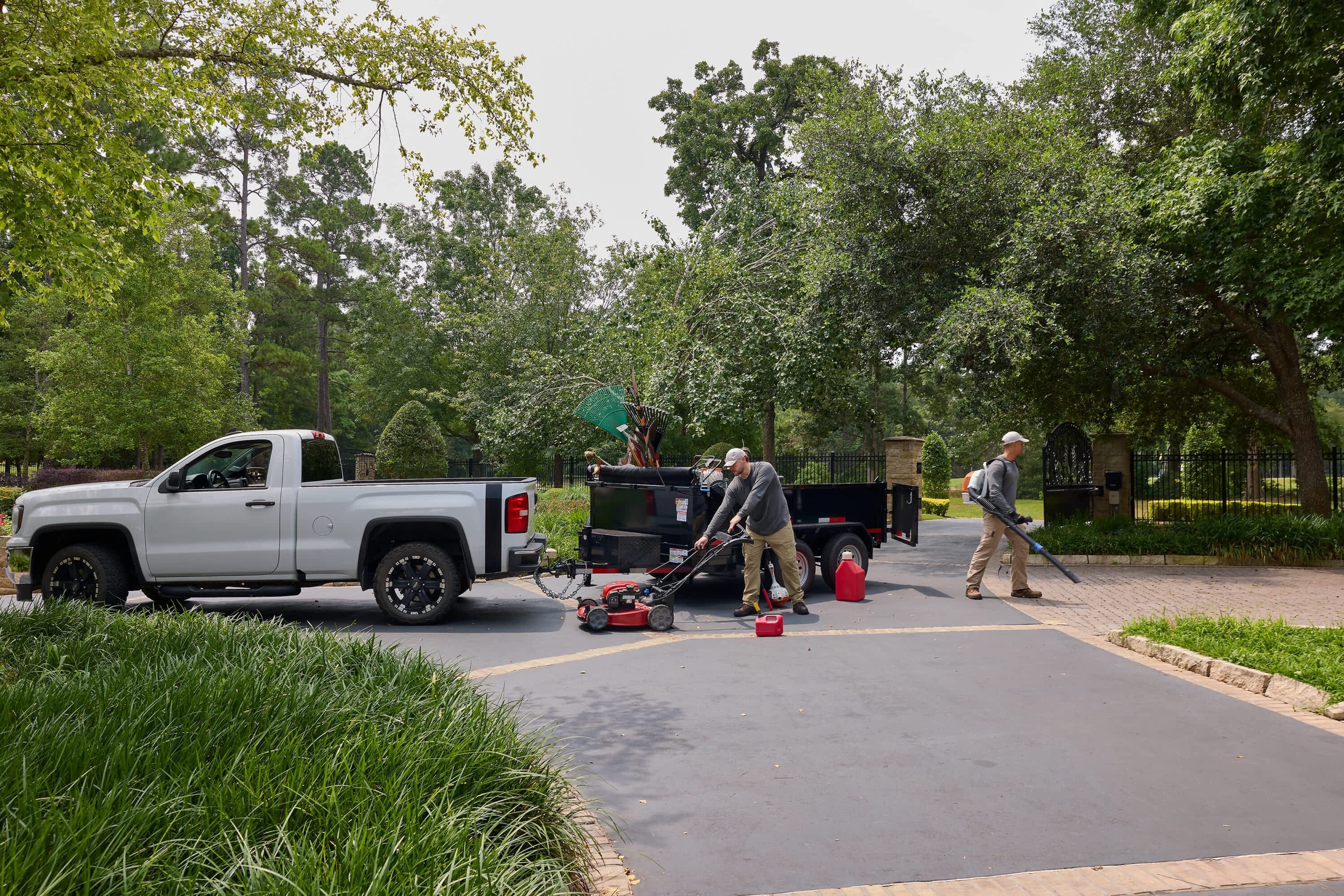 Two men do yard work, one with a mower and one with a leaf blower, near a Texas Pride Trailers dump trailer hitched to a white truck.
