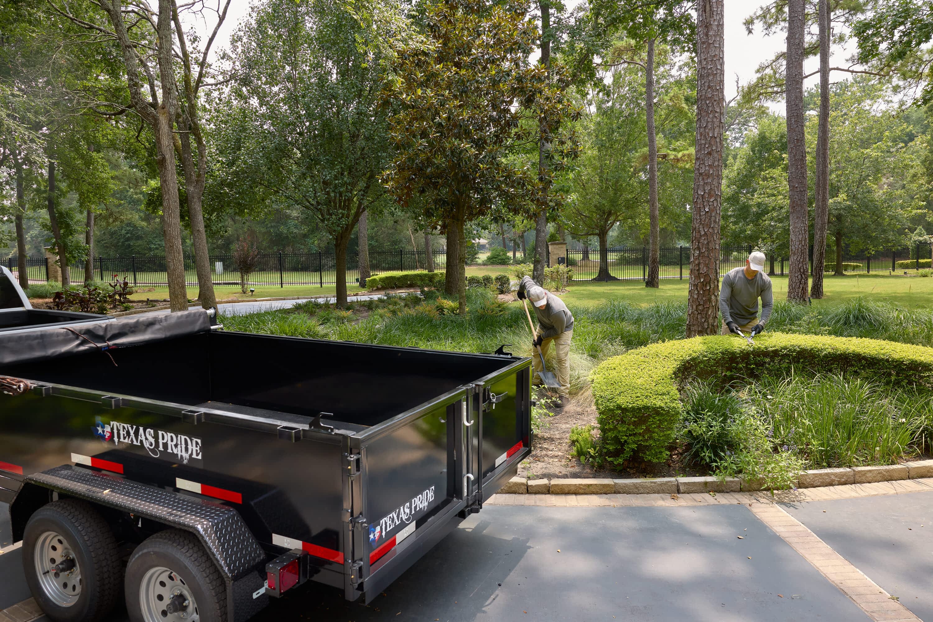 Two men do landscaping work, one digging and one trimming a hedge, near a Texas Pride Trailers dump trailer.