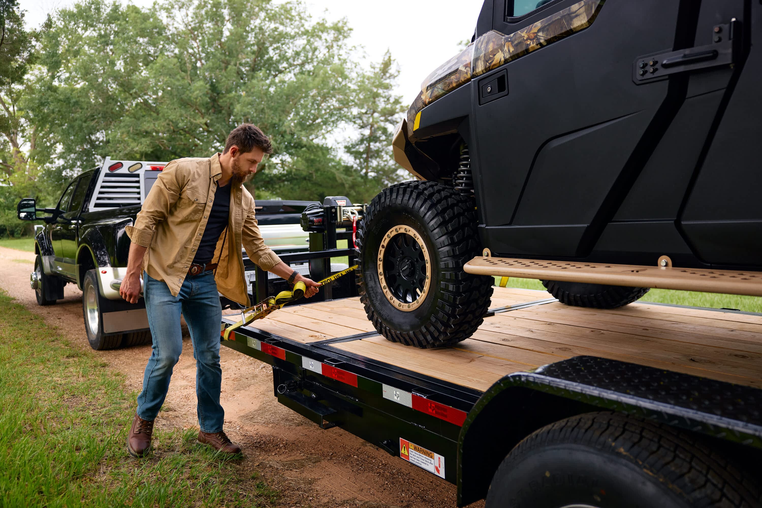 A man secures a UTV wheel to a Texas Pride Trailers flatbed trailer with a yellow strap. A black truck is in the background.