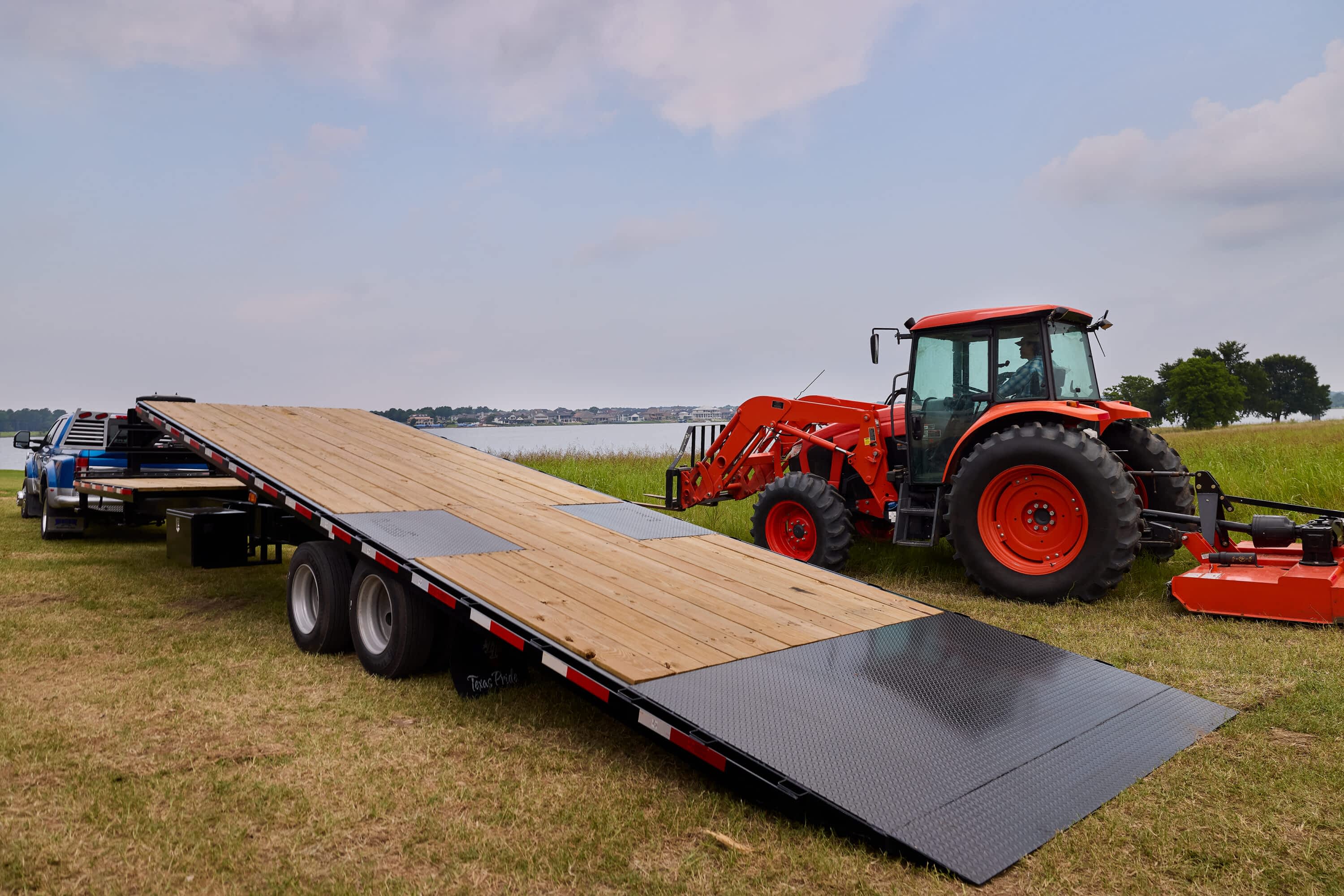 A Texas Pride Trailers flatbed trailer with a lowered hydraulic ramp sits in a grassy area near water. An orange tractor and brush hog are beside it, with a pickup truck attached to the front.