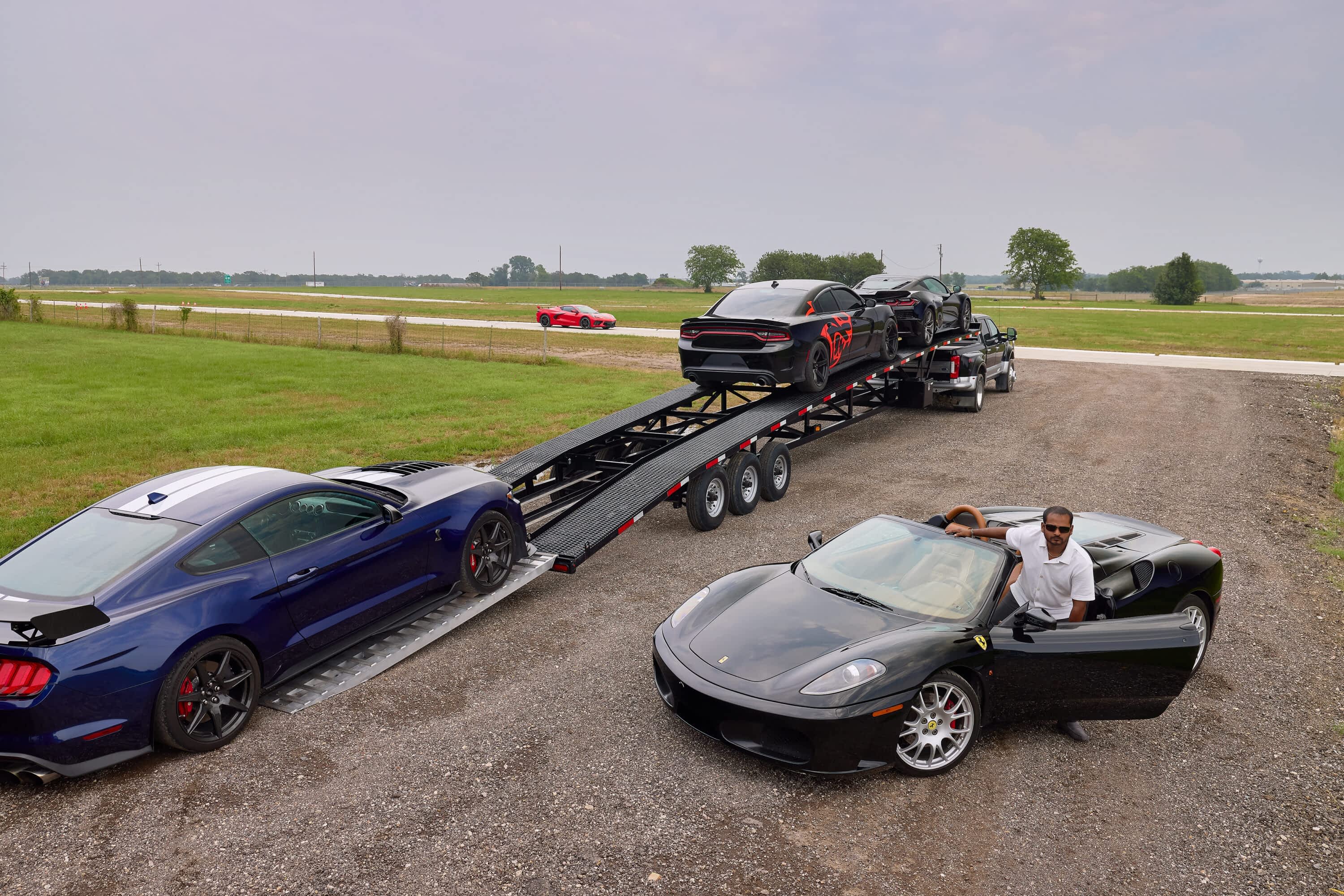 A man sits in a black convertible next to a Texas Pride Trailers multi-car hauler trailer loaded with a black and a blue car on a paved area, with a red car in the distance.