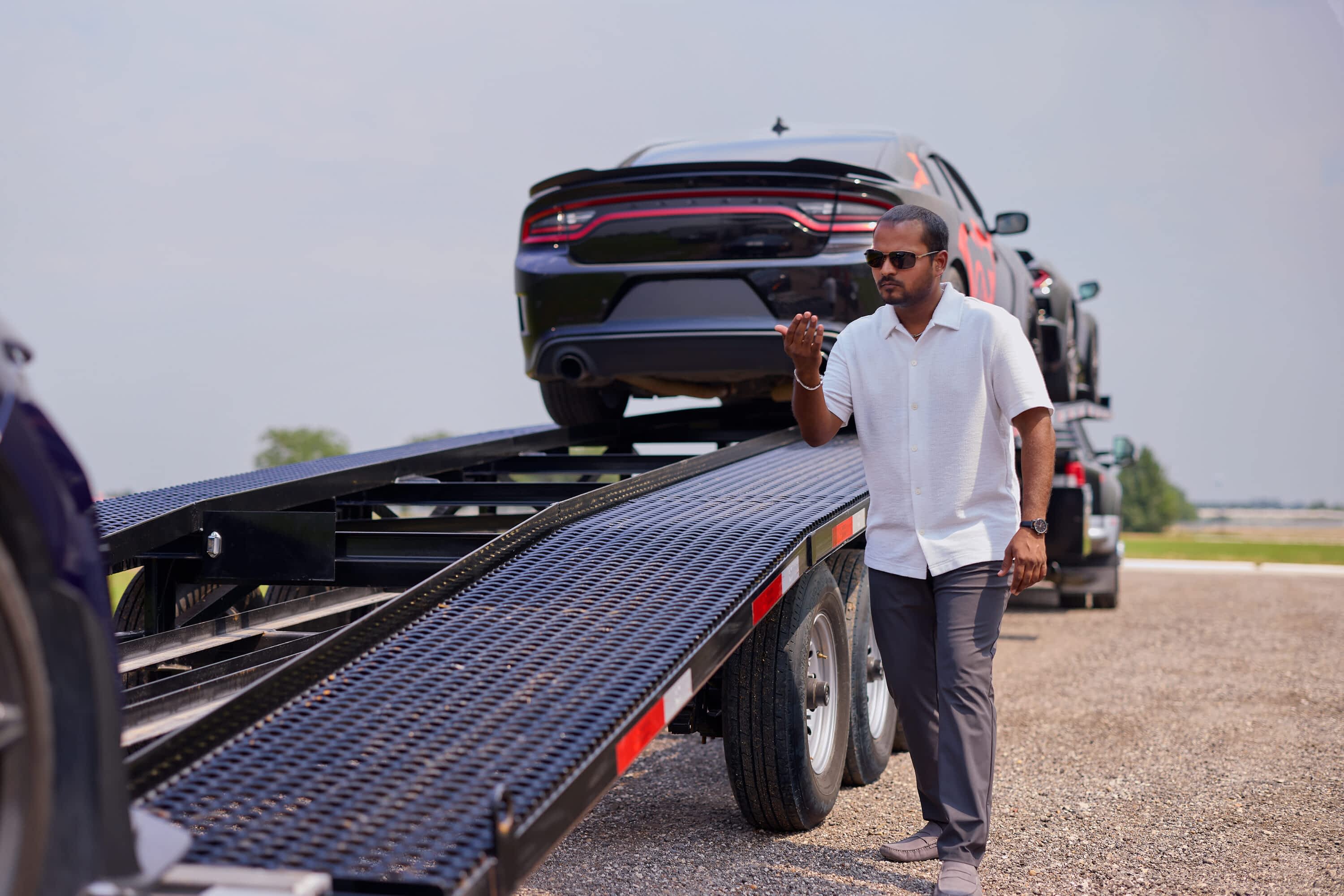 A man stands next to a Texas Pride Trailers multi-car hauler trailer with several black cars loaded on it, gesturing towards the trailer.