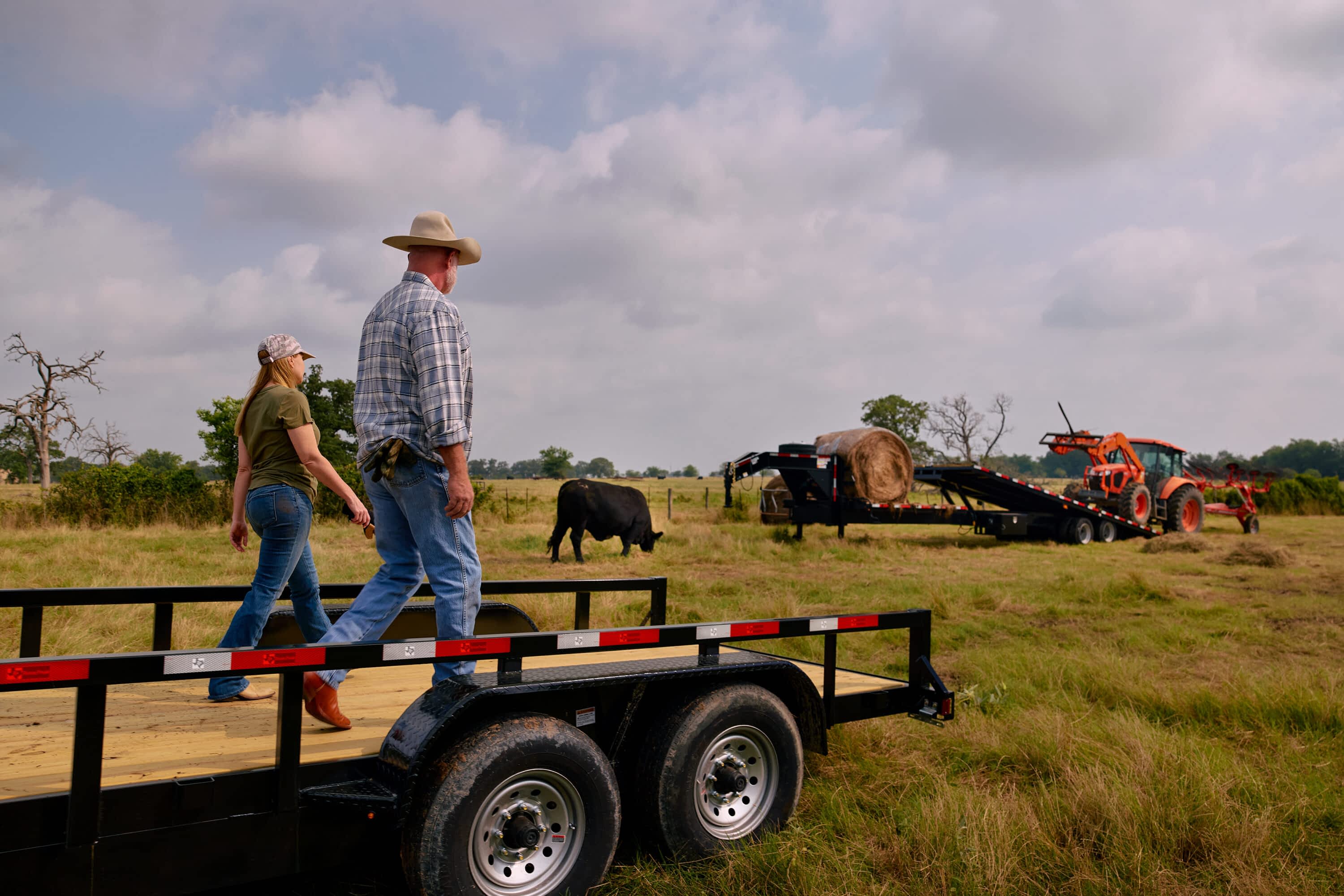A man and woman walk towards a Texas Pride Trailers flatbed trailer in a grassy field. Another Texas Pride Trailers trailer with a hay bale and a tractor is in the background with a cow grazing.
