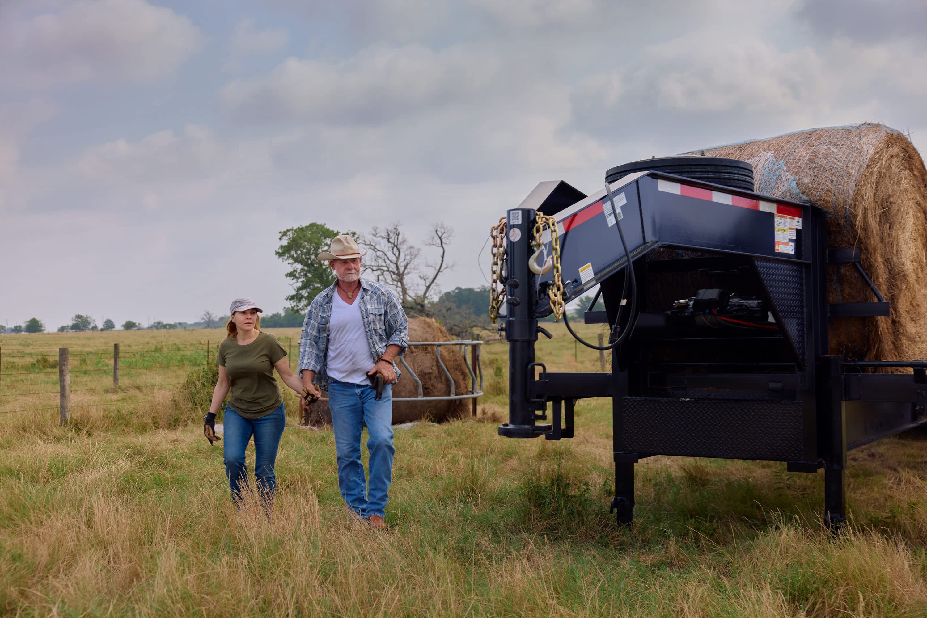 A man and woman walk through a grassy pasture beside a Texas Pride Trailers Flatbed 36K 8×40+5 Triple Axle Dual Wheel, which is loaded with large round hay bales. The trailer’s gooseneck hitch, safety chains, and hydraulic components are visible in the foreground, with a wire fence, trees, and overcast sky in the background.