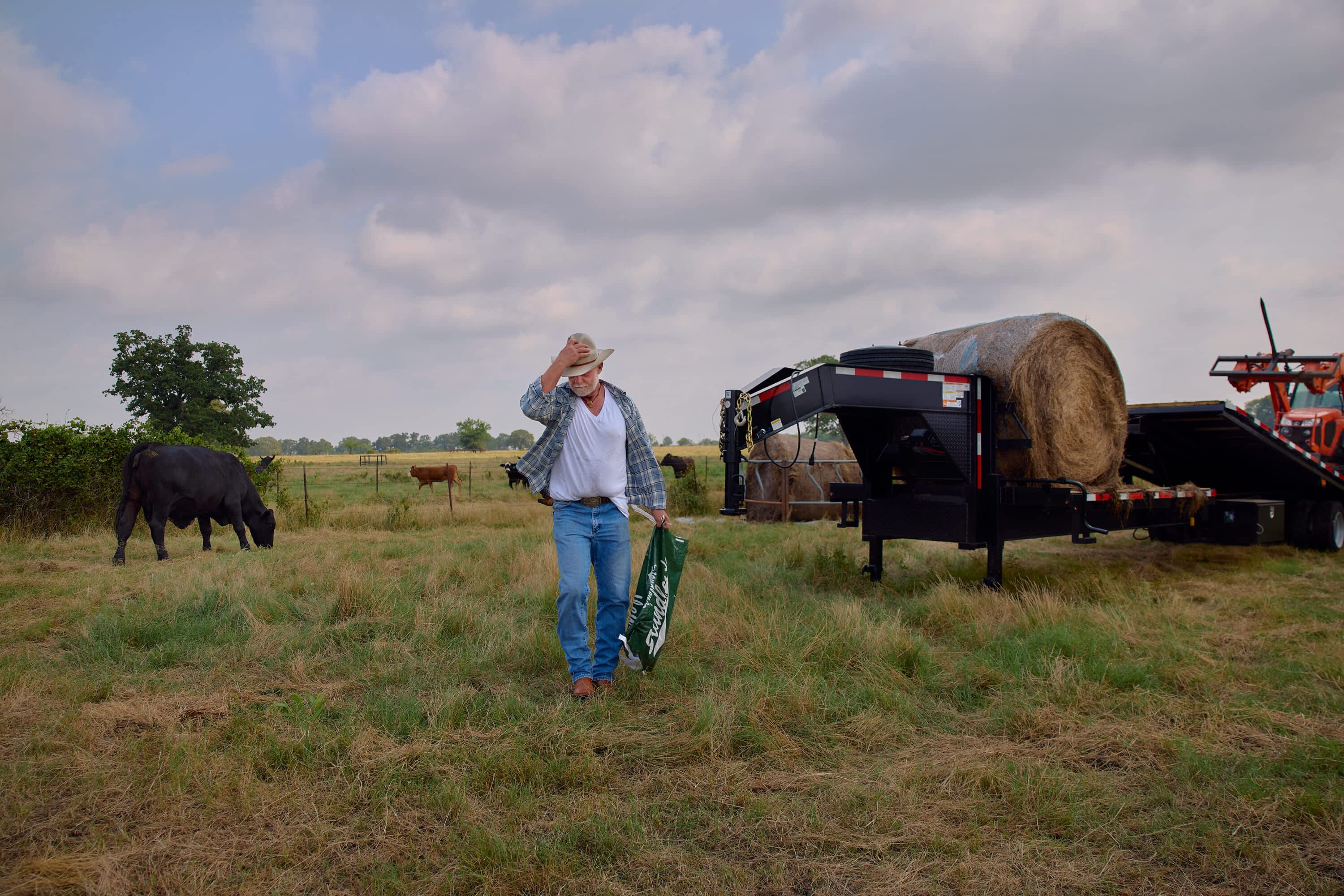 A man walks across a grassy field carrying a bag, with a Texas Pride Trailers gooseneck trailer loaded with hay bales and a tractor behind him, and cattle grazing in the distance.