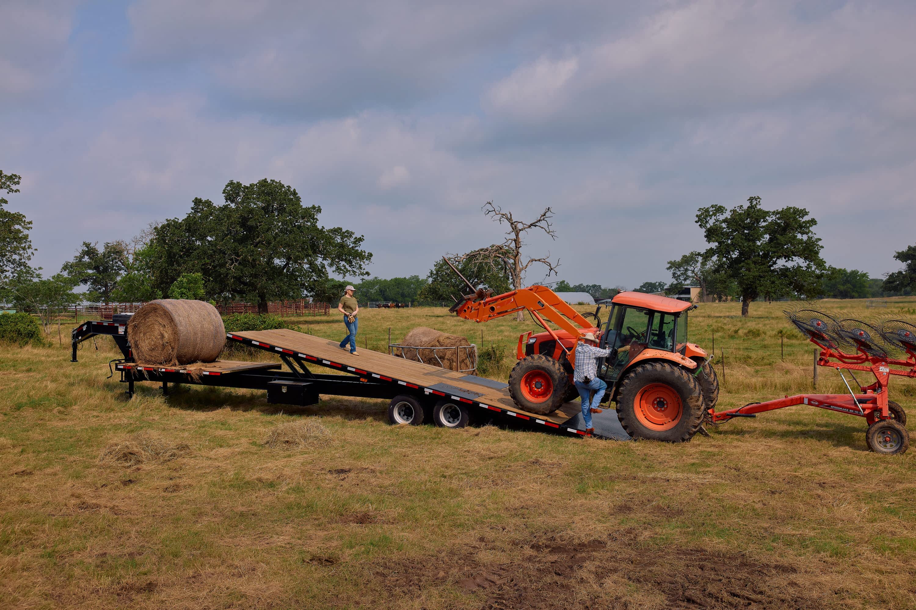 Two people stand near a hay bale and an orange Kubota tractor with a plow on a Texas Pride Trailers flatbed trailer in a grassy field.
