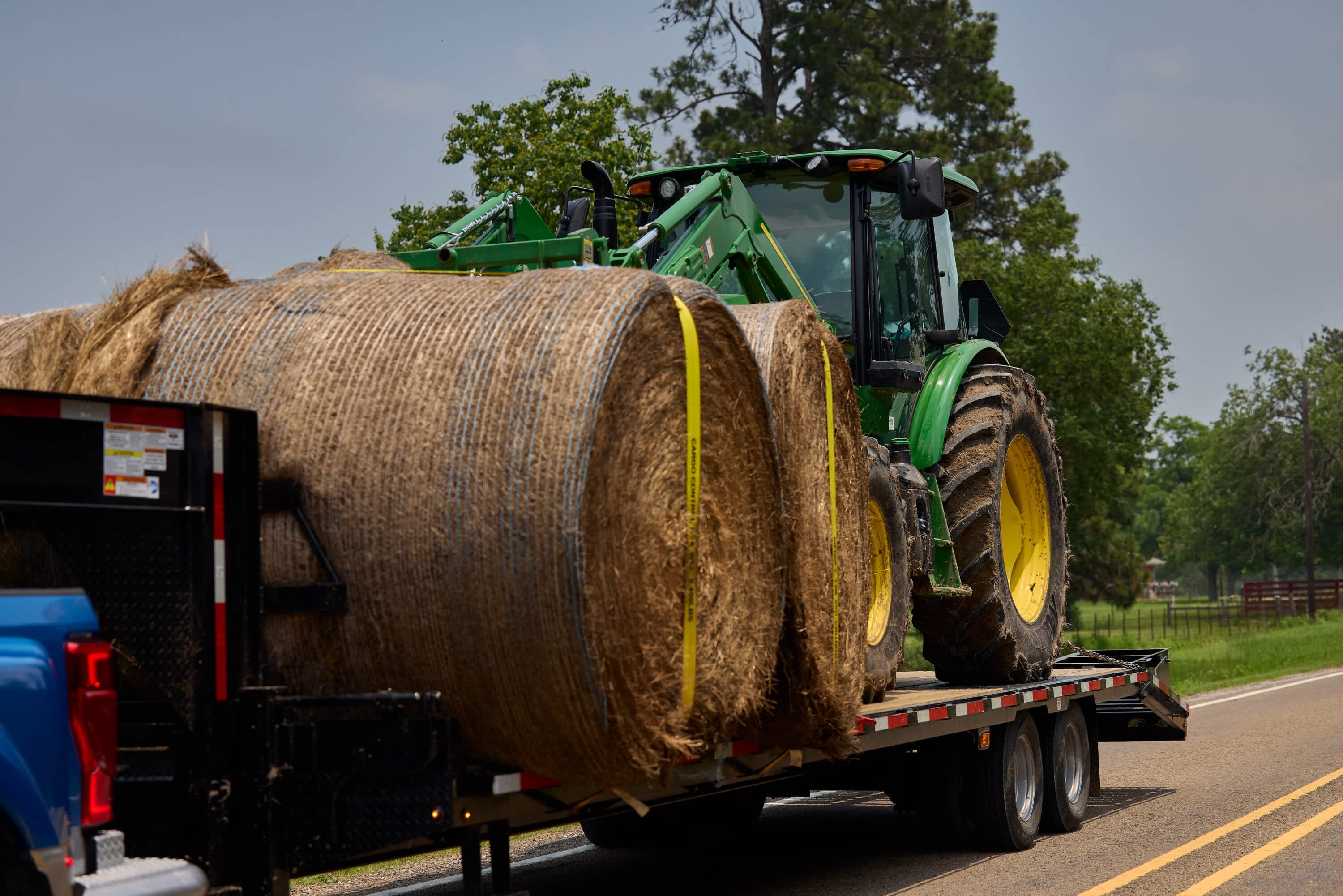 A Texas Pride Trailers Flatbed 26K 8×25+5 Gooseneck Dual Wheel is loaded with large round hay bales and a green John Deere tractor, secured with yellow straps. The trailer is hitched to a blue pickup truck and traveling down a rural two-lane road lined with trees and fencing under bright afternoon light.