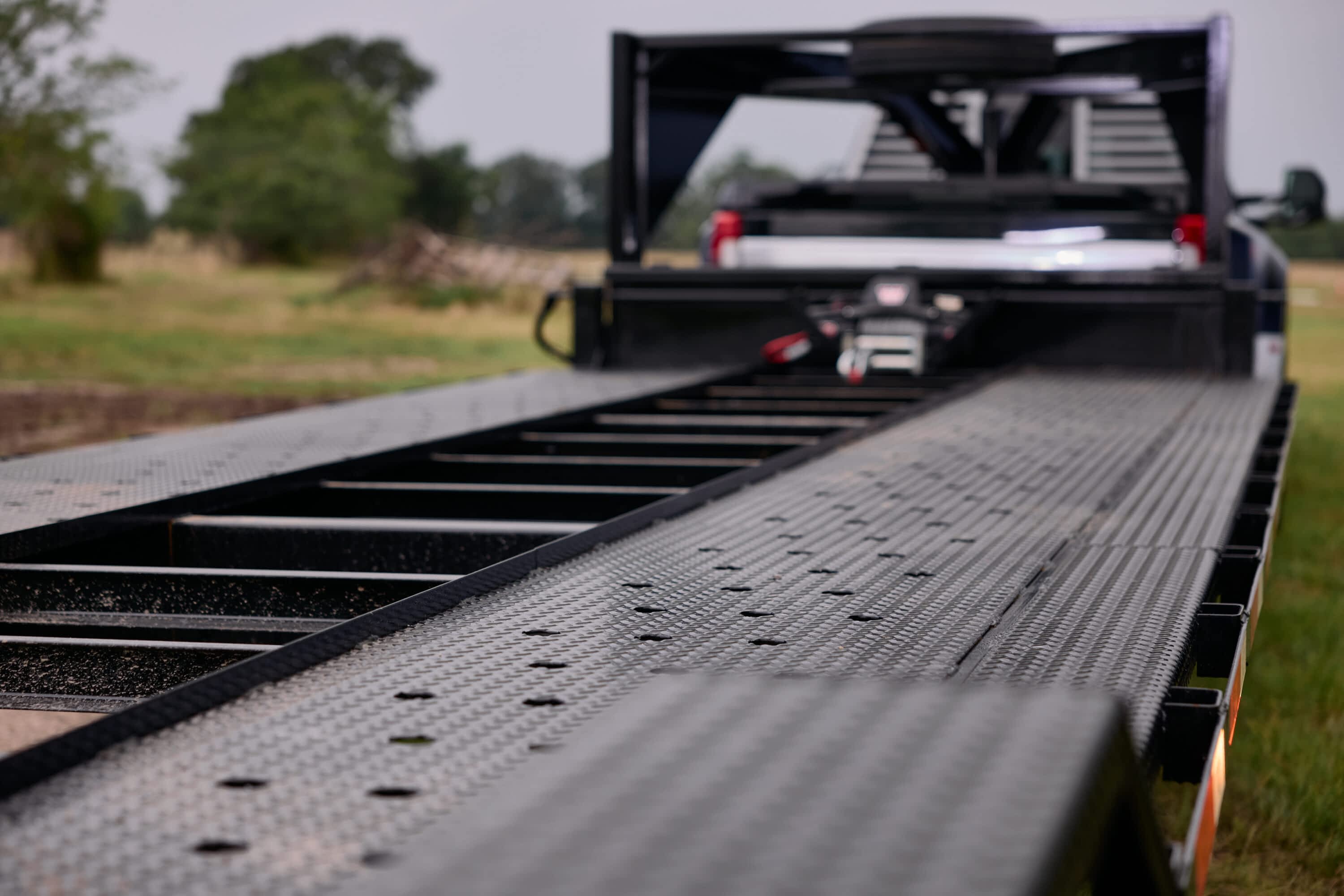 Close-up view of a Texas Pride 2-car hauler trailer with drive-over fenders and a perforated diamond plate steel deck. The trailer is hitched to a pickup truck and parked in a grassy field, highlighting its rugged construction and vehicle loading capabilities.