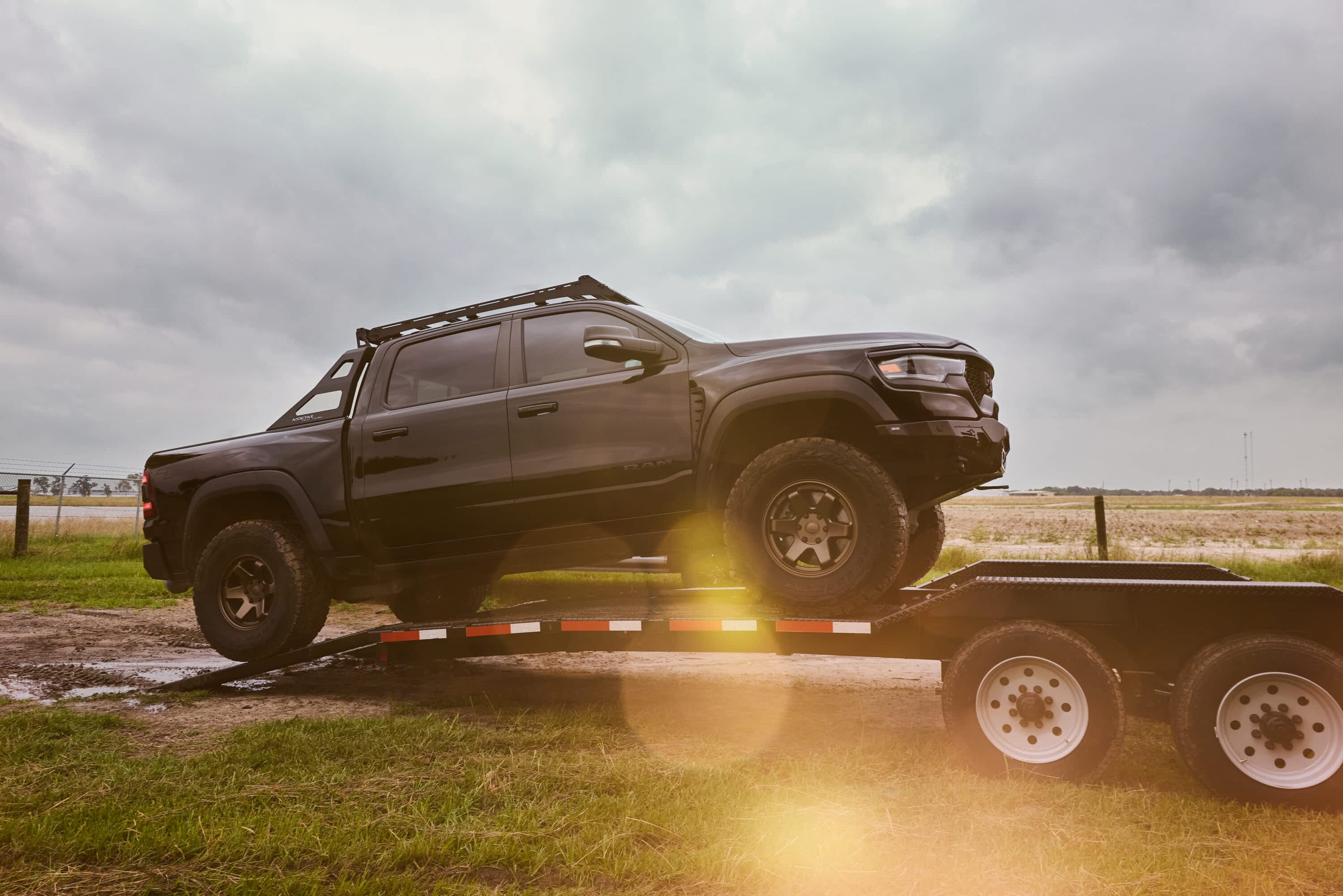 A black off-road pickup truck is being loaded onto a Texas Pride car hauler trailer in a grassy field under cloudy skies, with sunlight flaring through the camera lens.