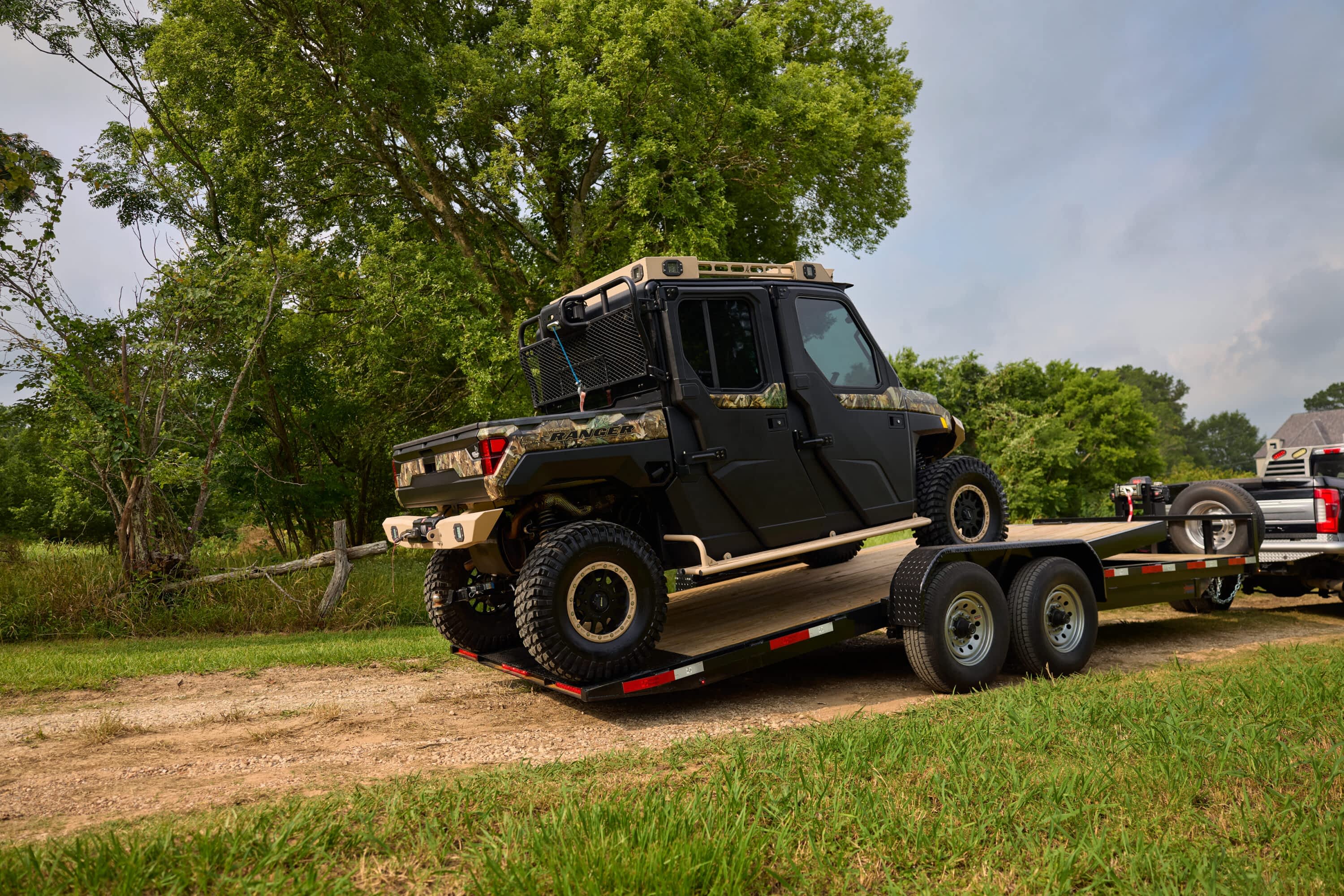 A tan-and-black side-by-side off-road vehicle sits loaded on a Texas Pride gravity tilt trailer hitched to a pickup truck. The trailer is parked on a dirt path surrounded by green grass and trees, showcasing easy equipment transport in a rural setting.