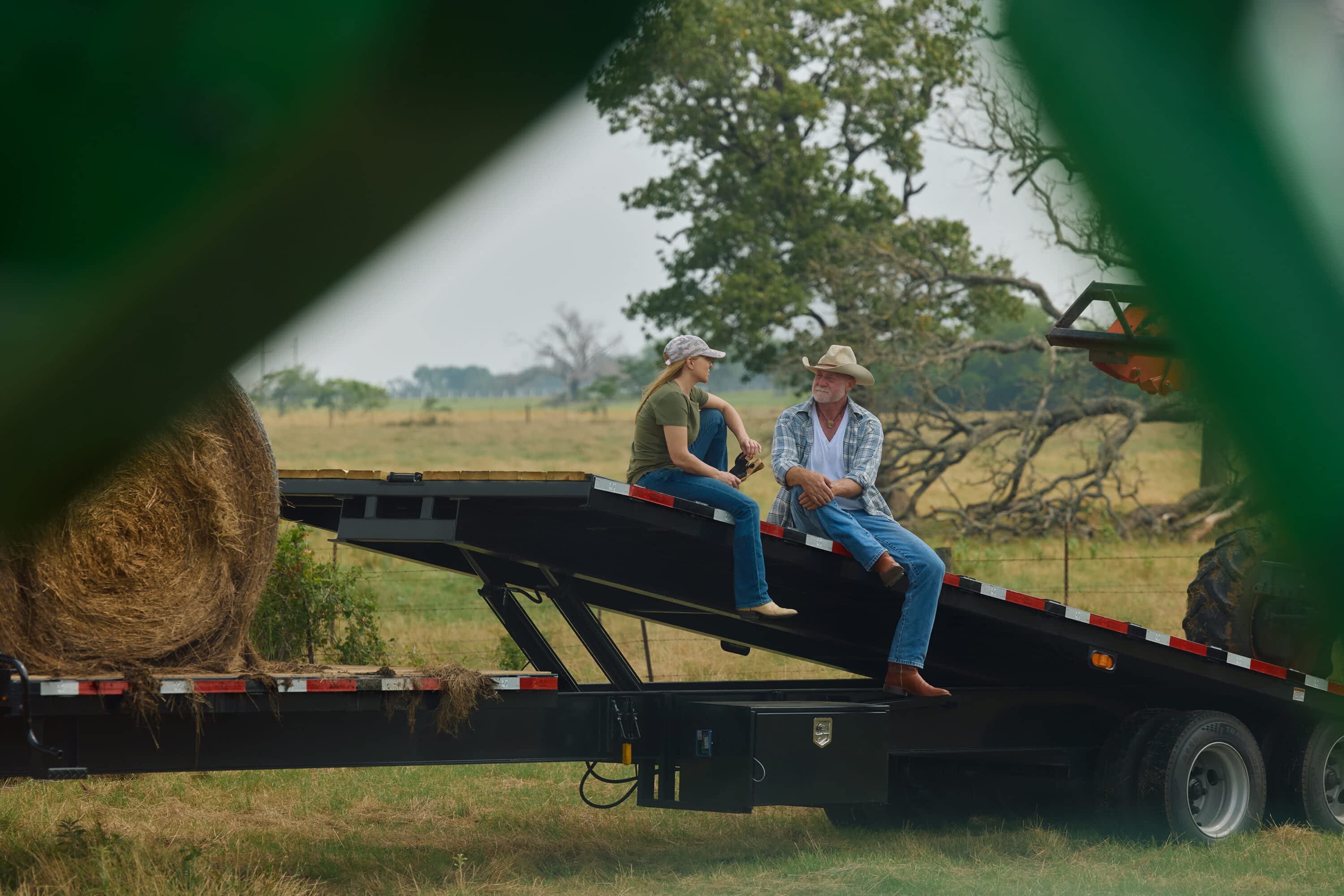 A man and woman sit on the tilted deck of a Texas Pride power tilt trailer in a rural pasture, with a large hay bale loaded on the trailer and a tractor nearby. The relaxed moment captures the versatility of the trailer for both heavy lifting and everyday ranch life.