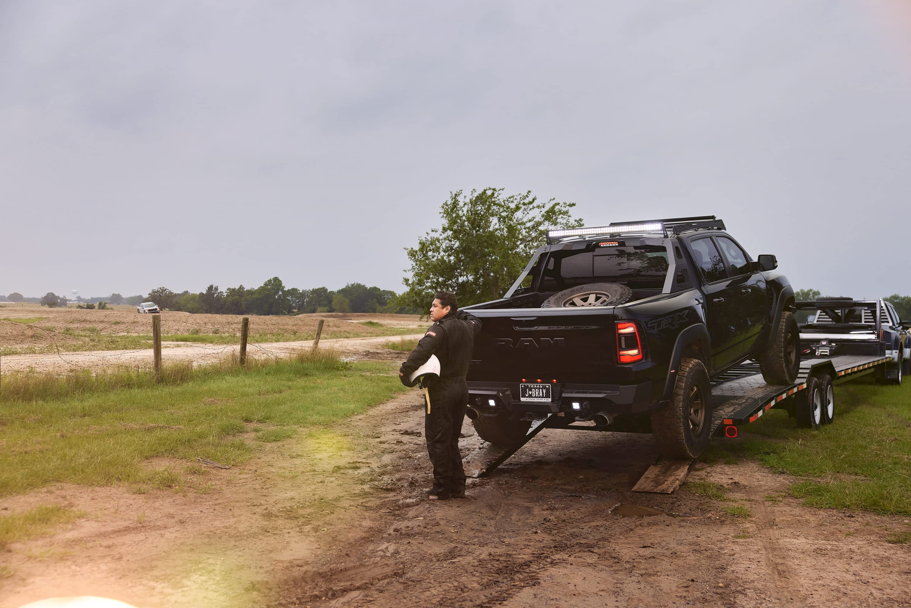A person stands next to a black Ram truck being loaded onto a Texas Pride Trailers car hauler trailer on a dirt road in a field.