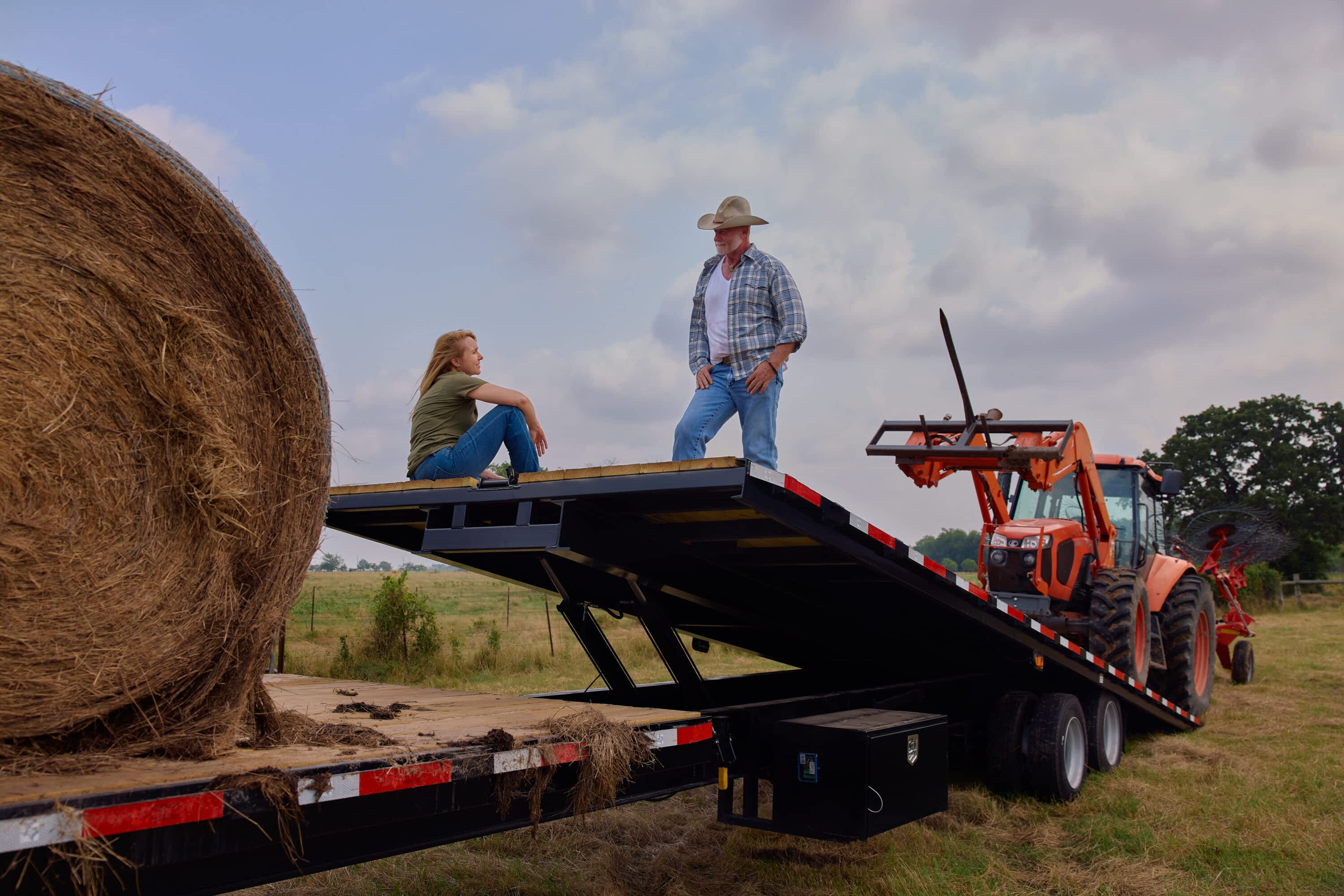 Texas Pride triple-axle gooseneck power tilt trailer in a rural field, with a large round hay bale on the deck and an orange tractor with a hay rake attachment loaded on the raised tilt bed. A man in a cowboy hat stands on the tilted deck while a woman sits beside him, both facing each other, framed by open pasture and cloudy skies.