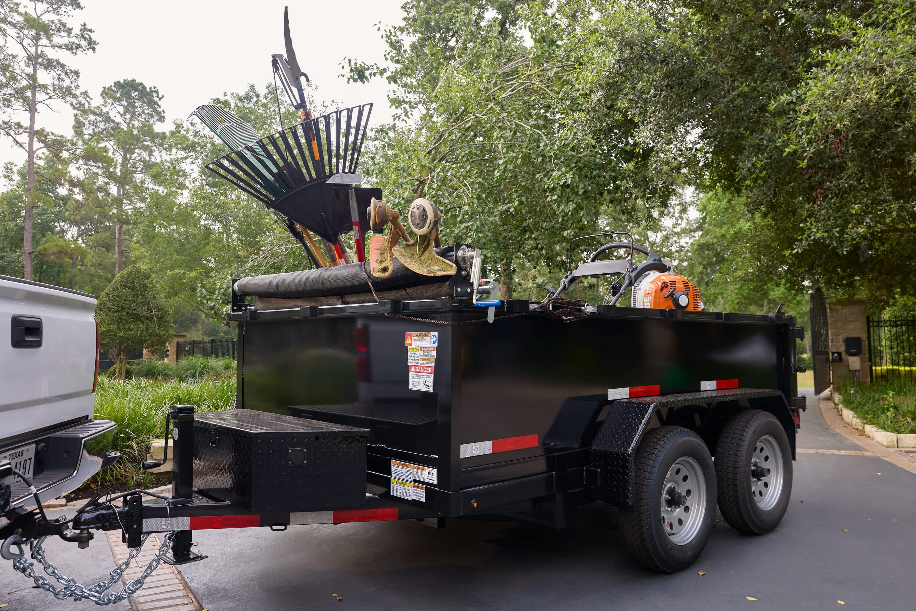 A Texas Pride Trailers dump trailer loaded with yard waste and equipment is hitched to a white truck and parked on a paved area.