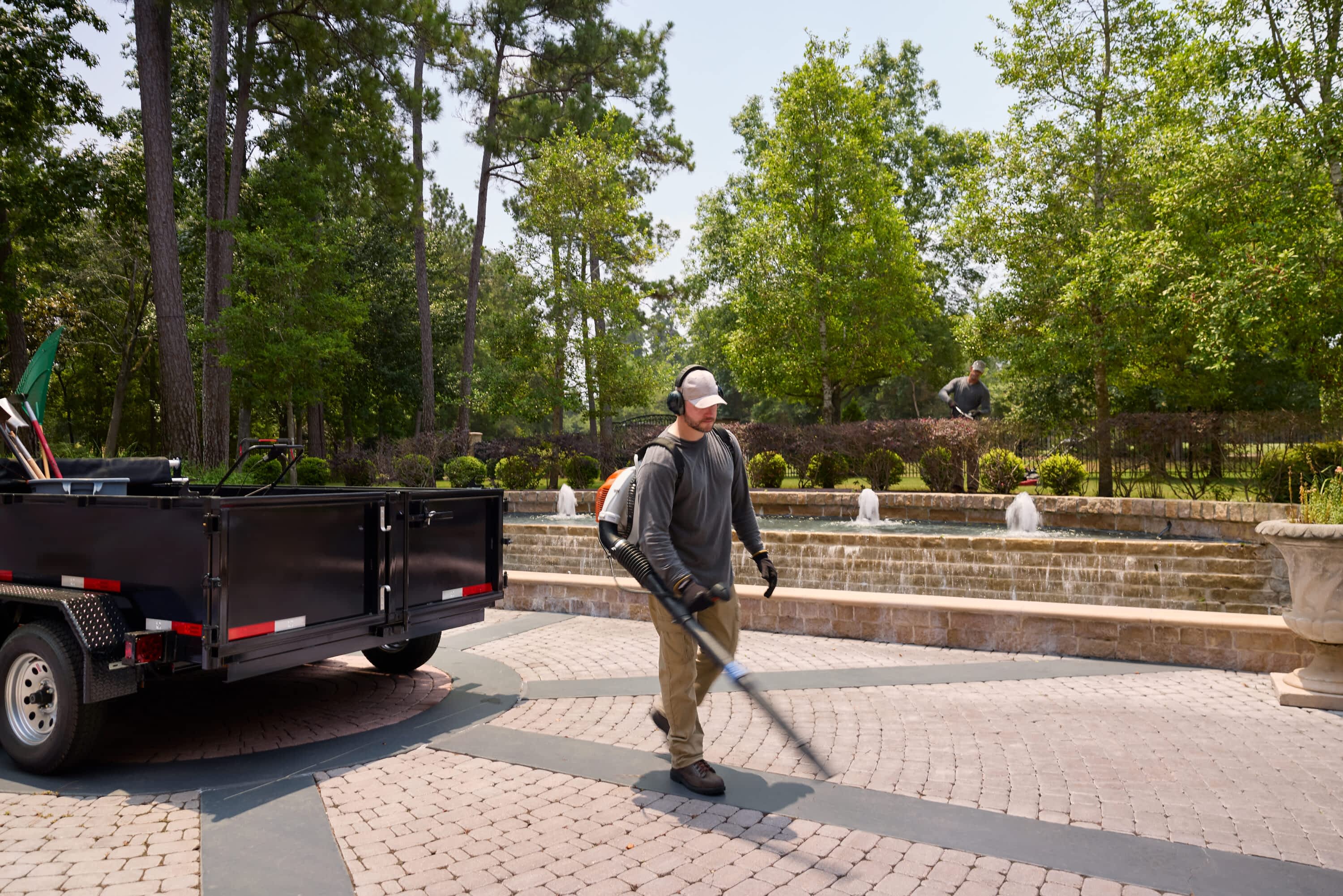 A man uses a leaf blower on a paved area near a Texas Pride Trailers dump trailer loaded with tools. There is a fountain and landscaping in the background.