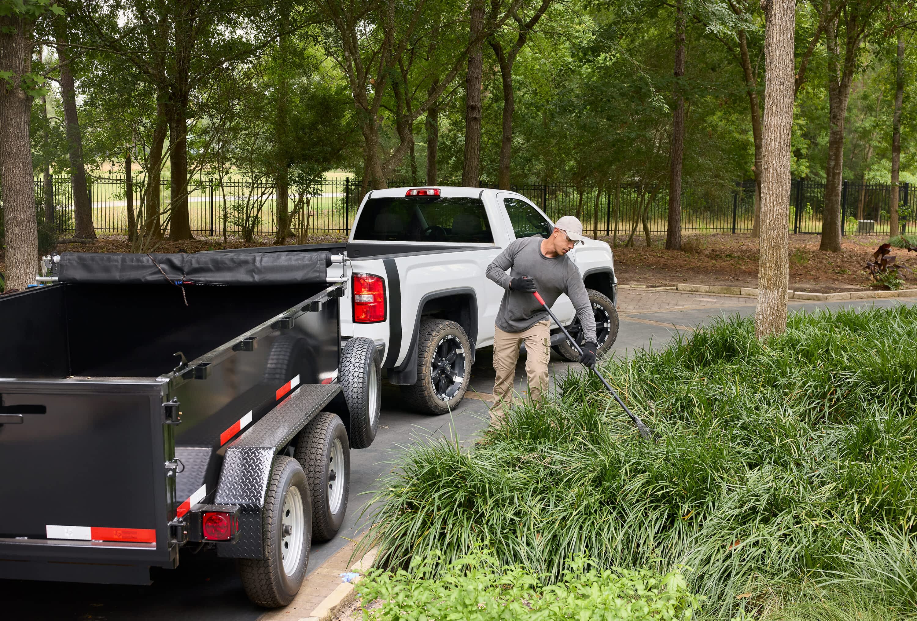 A man rakes debris from a hedge next to a Texas Pride Trailers dump trailer hitched to a white truck.