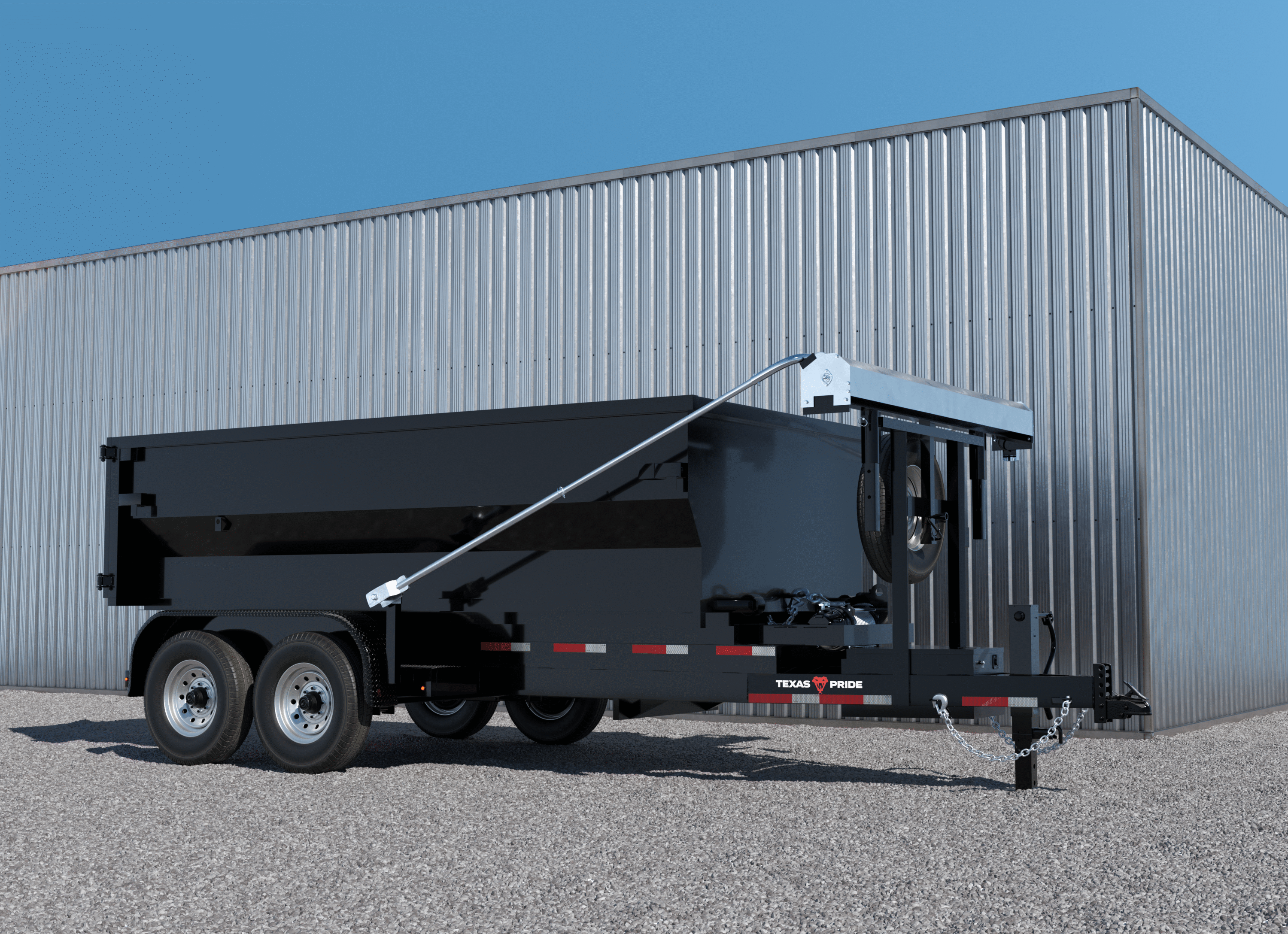 Black Texas Pride bumper pull roll-off trailer with a loaded dumpster and extended tarp arm, shown parked on gravel in front of a metal building—illustrating the process of loading your dumpster onto your Texas Pride roll-off.