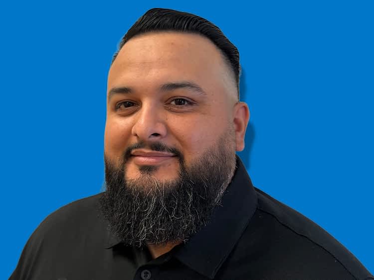 Texas Pride Trailers Location Manager in Oakridge, Oregon with short gray hair, glasses, and a mustache, wearing a black T-shirt in front of a blue background.