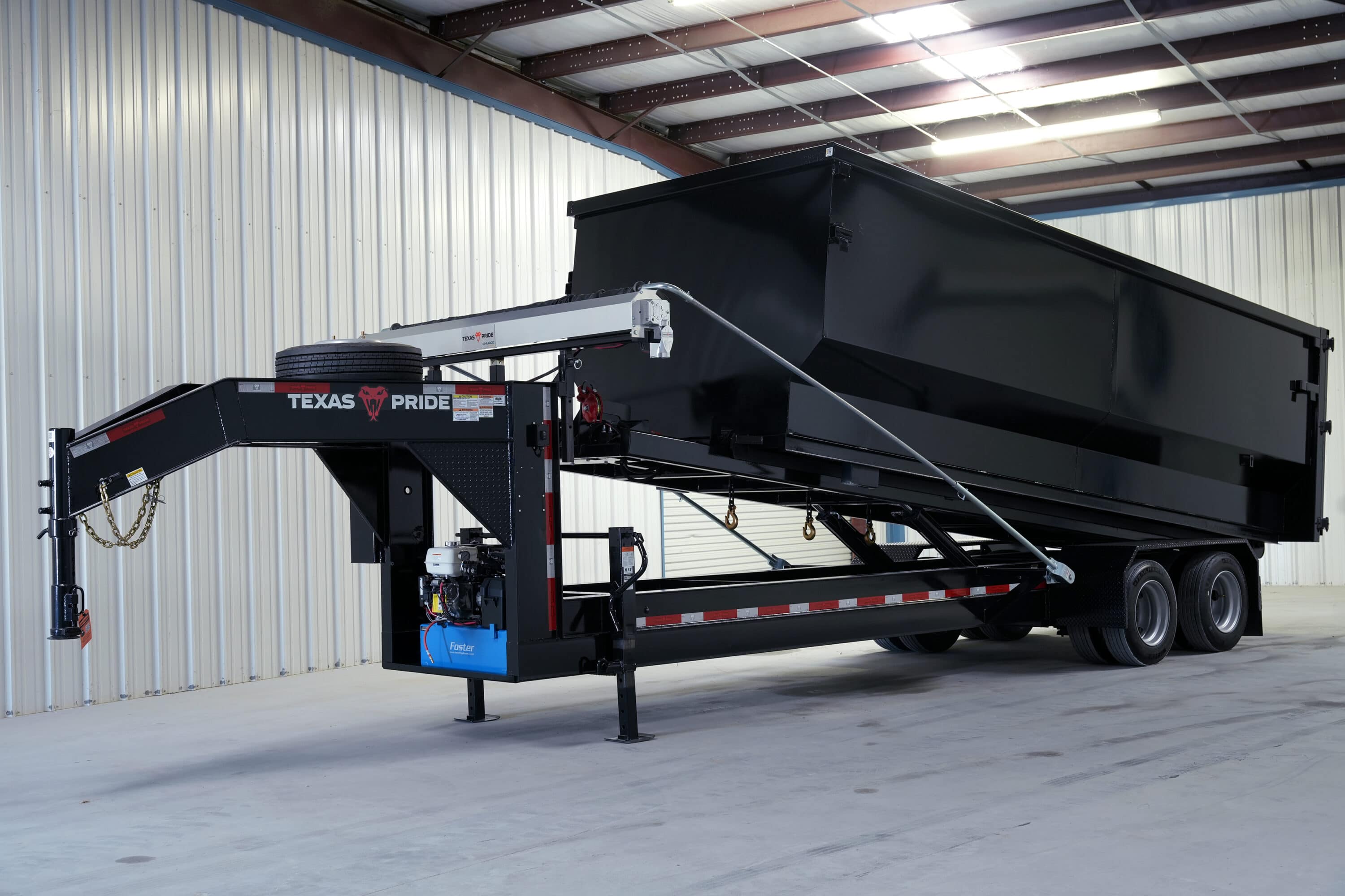 Black Texas Pride gooseneck roll-off trailer with a large black container raised at an angle, showing a gas engine and blue hydraulic reservoir on the gooseneck, a spare tire mounted above it, and dual tandem wheels, in a warehouse.
