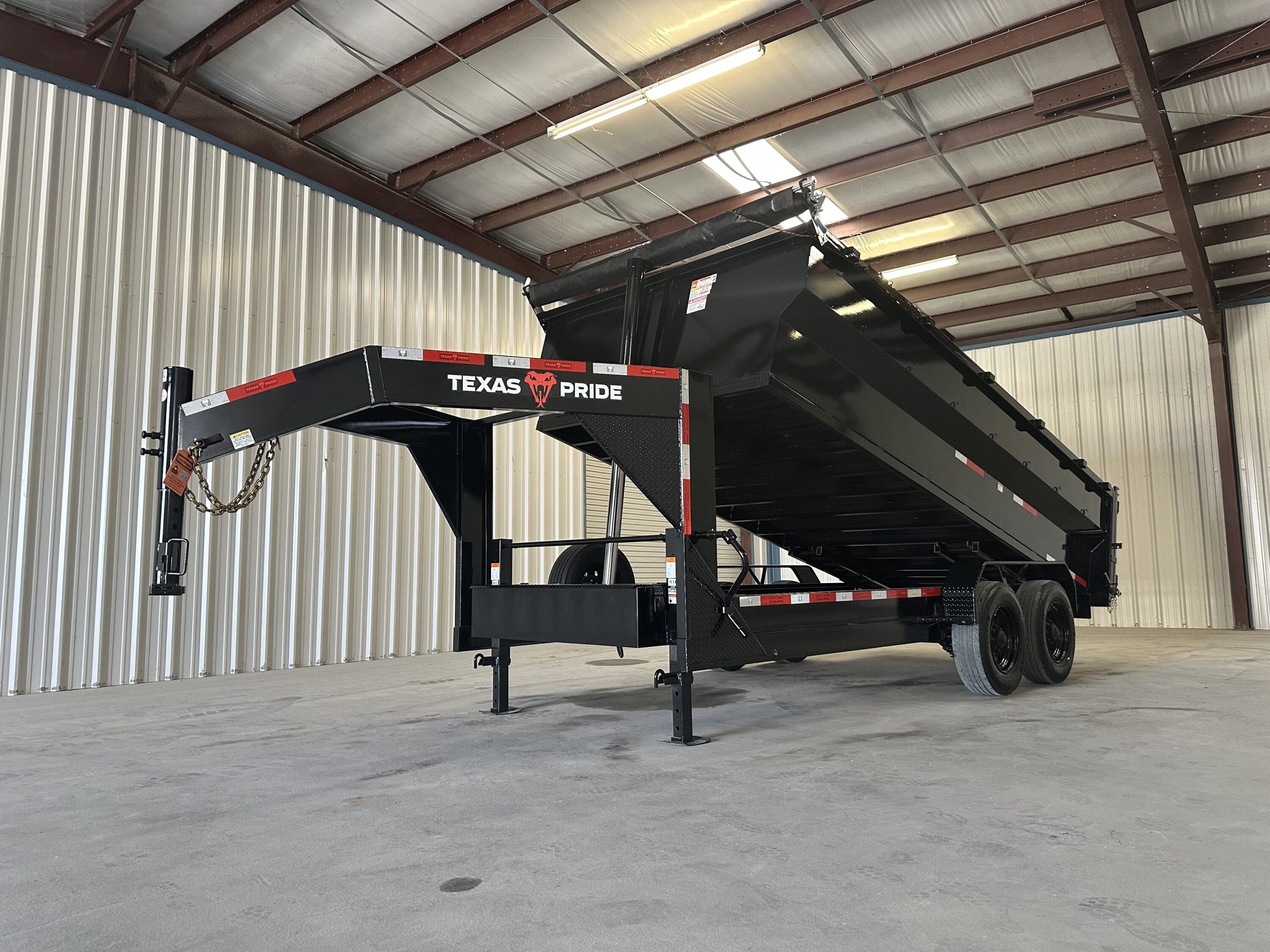 Black Texas Pride gooseneck dump trailer with the bed raised in the dump position and a rolled tarp at the top, standing on tandem black wheels and front stabilizer jacks inside a warehouse.